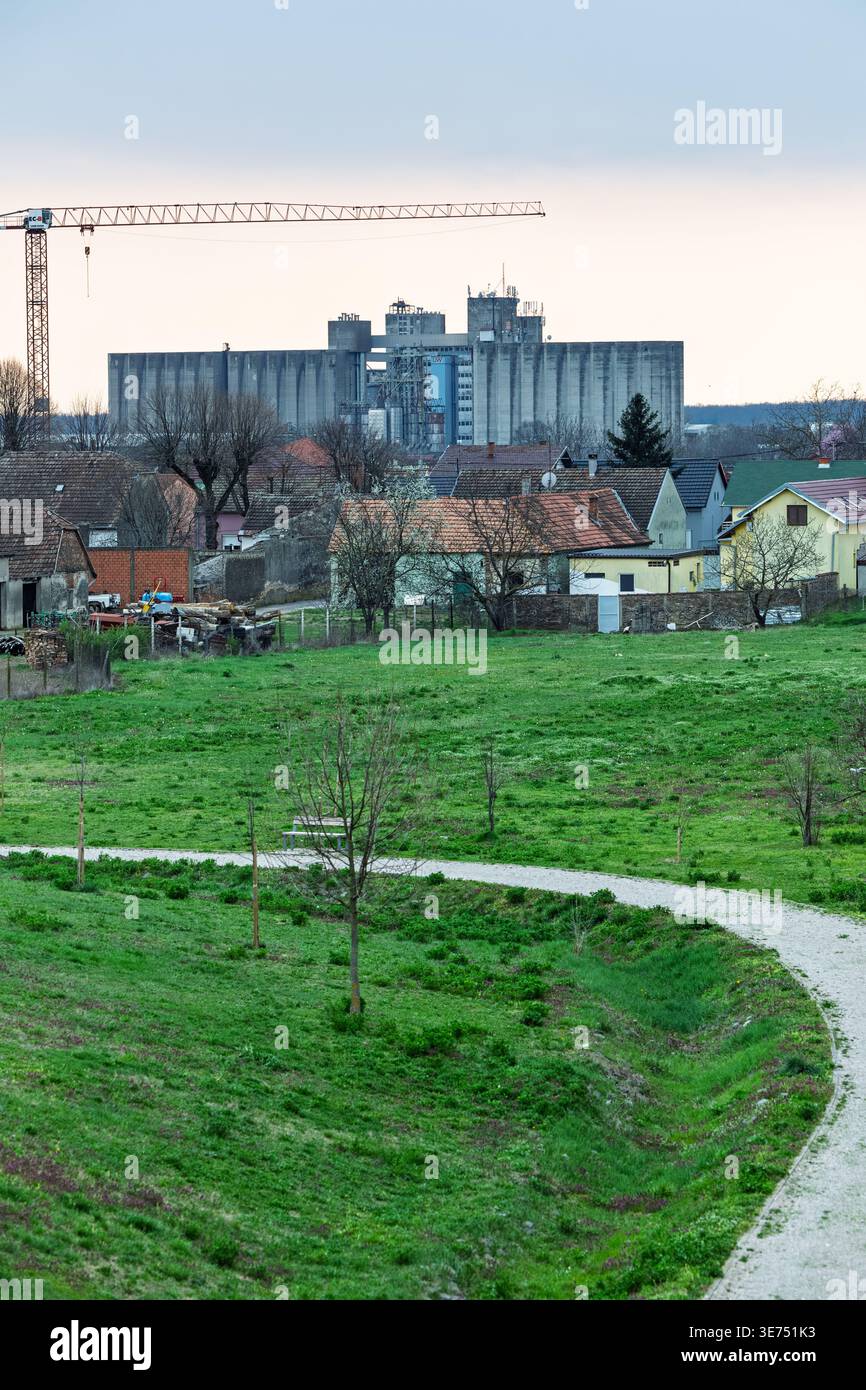 Ein ruhiges ländliches Dorf mit Ziegeldächern und einem gewundenen Pfad im Vordergrund, dominiert von großen Betonkornsilos und einem Baukran auf der h Stockfoto