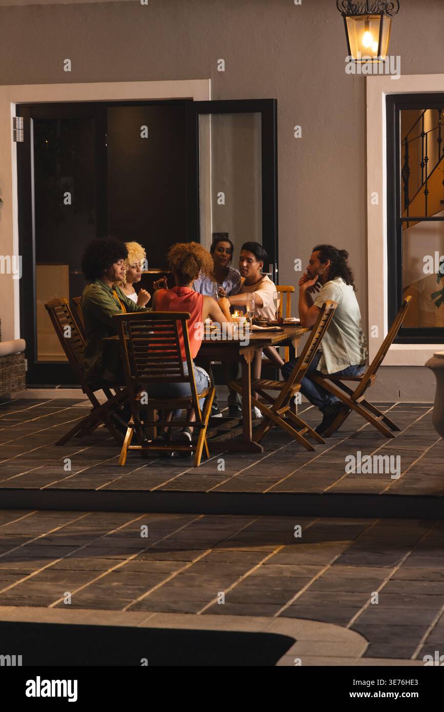 Verschiedene Freunde, die legere Kleidung tragen, lehnen sich hinein und teilen sich das Essen am Holztisch auf der gefliesten Terrasse Stockfoto