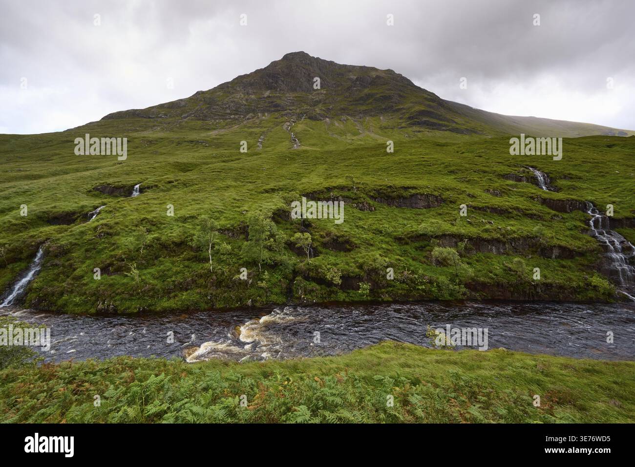 Üppiges Grün mit kleinen Wasserfällen und einem Berg im Hintergrund unter einer bewölkten Decke, Glen Etive, Schottland, Großbritannien Stockfoto