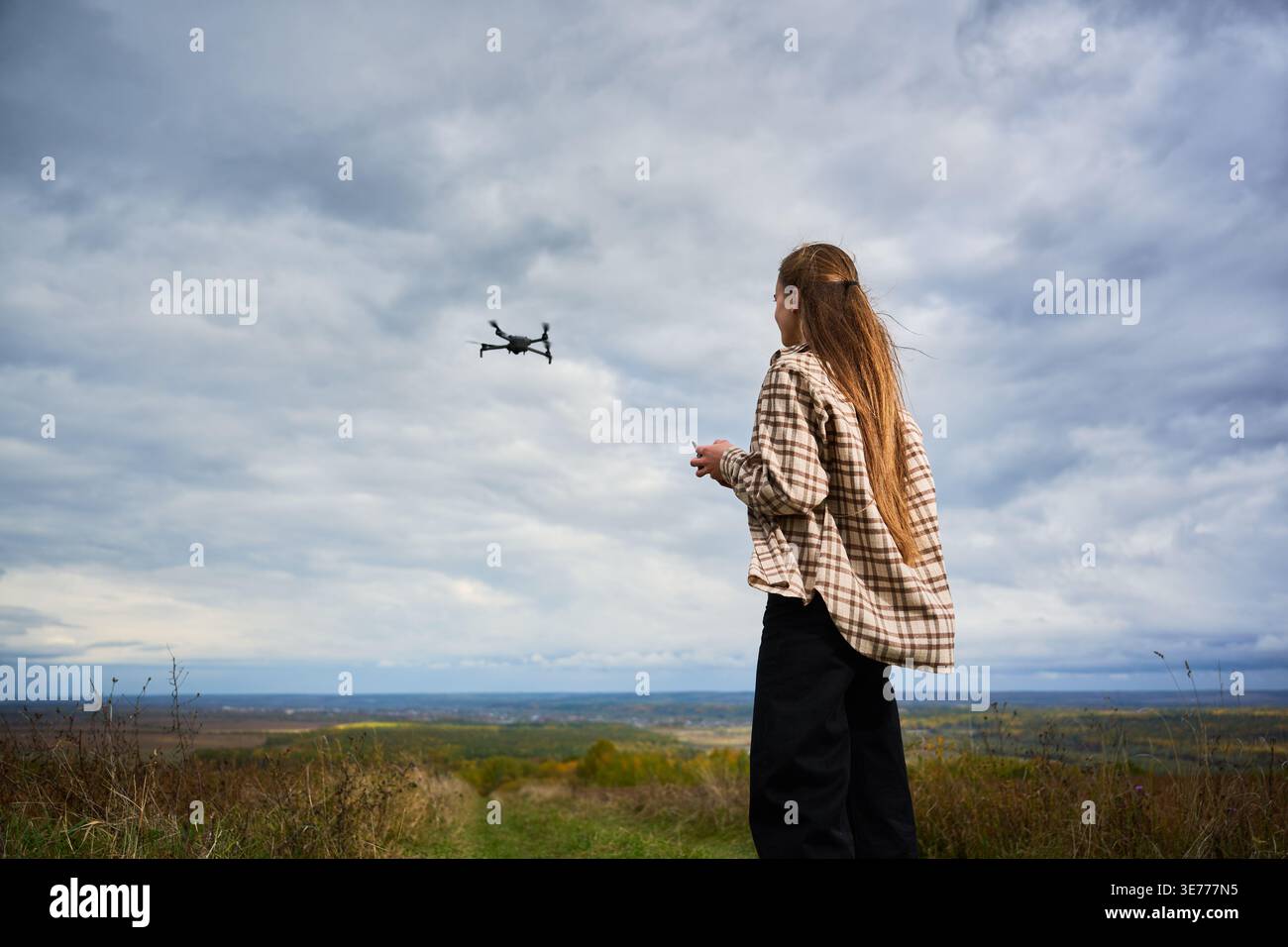 Rückansicht einer Frau, die Drohne kontrolliert, die bei bewölktem Himmel über weite Landschaft fliegt. Pilotin mit Fernbedienung auf dem Hügel, Luftaufnahmen und Explorationskonzept der Technologie Stockfoto