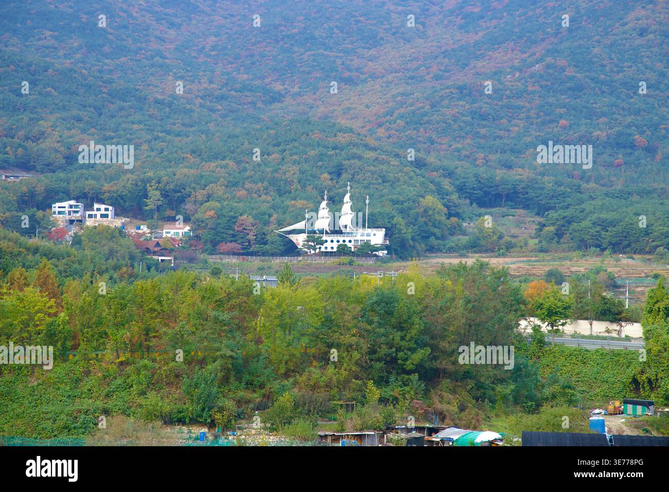 In Ulju-Gun, Ulsan, erscheint ein schiffförmiges weißes Gebäude quer durch das Tal, eingerahmt von bewaldeten Hängen, verstreuten ländlichen Häusern und gedämpften Herbstfarben. Stockfoto