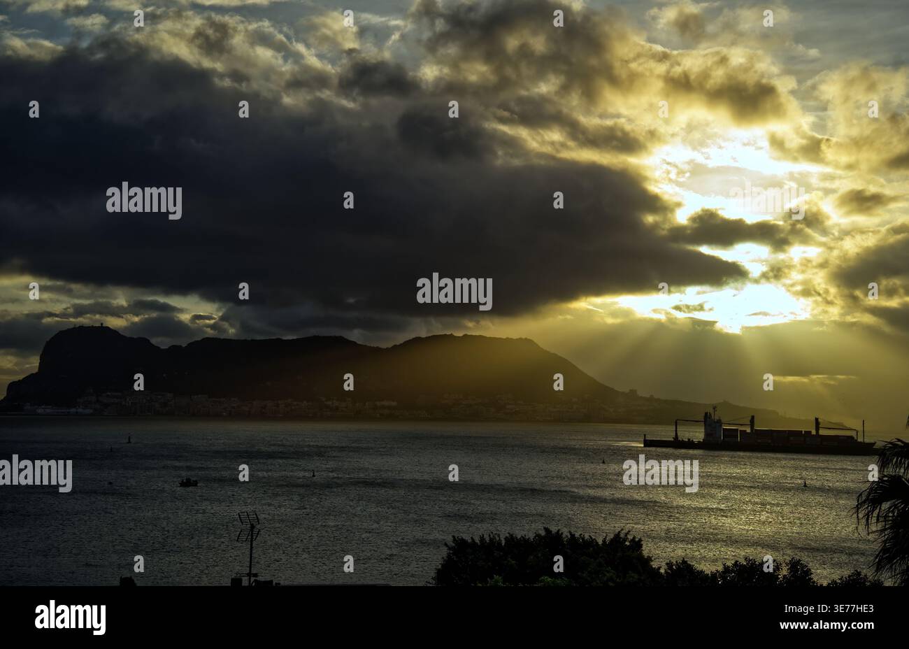 Das goldene Licht der Sonne bricht durch dunkle, schwere Wolken und wirft dramatische Strahlen über die Silhouette des Felsens von gibraltar und das ruhige mittelmeer Stockfoto
