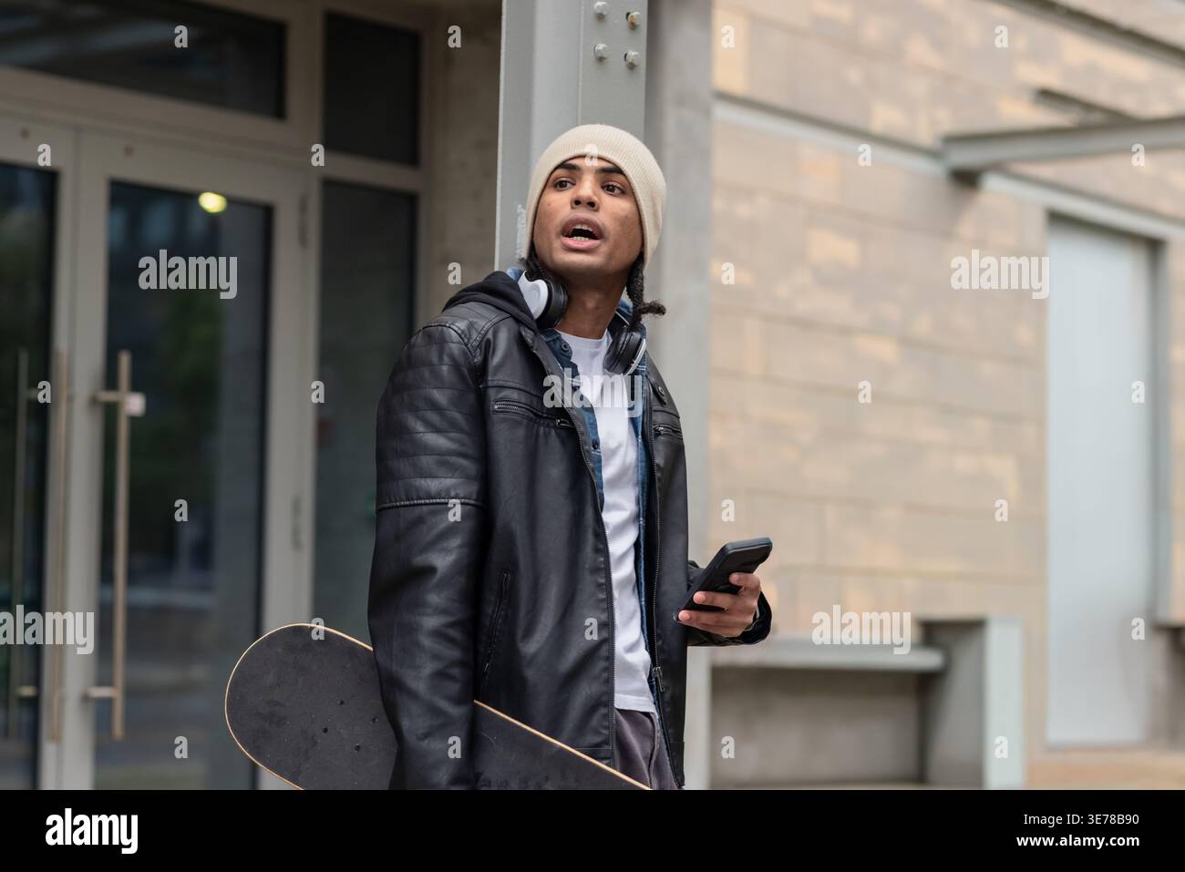 Mann, der sich am Eingang gegen die Säule lehnt, trägt eine Mütze, die Skateboard hält und das Smartphone überprüft. Urban, Outdoor, Lifestyle, Kopfhörer, Telefon, Skate, jac Stockfoto