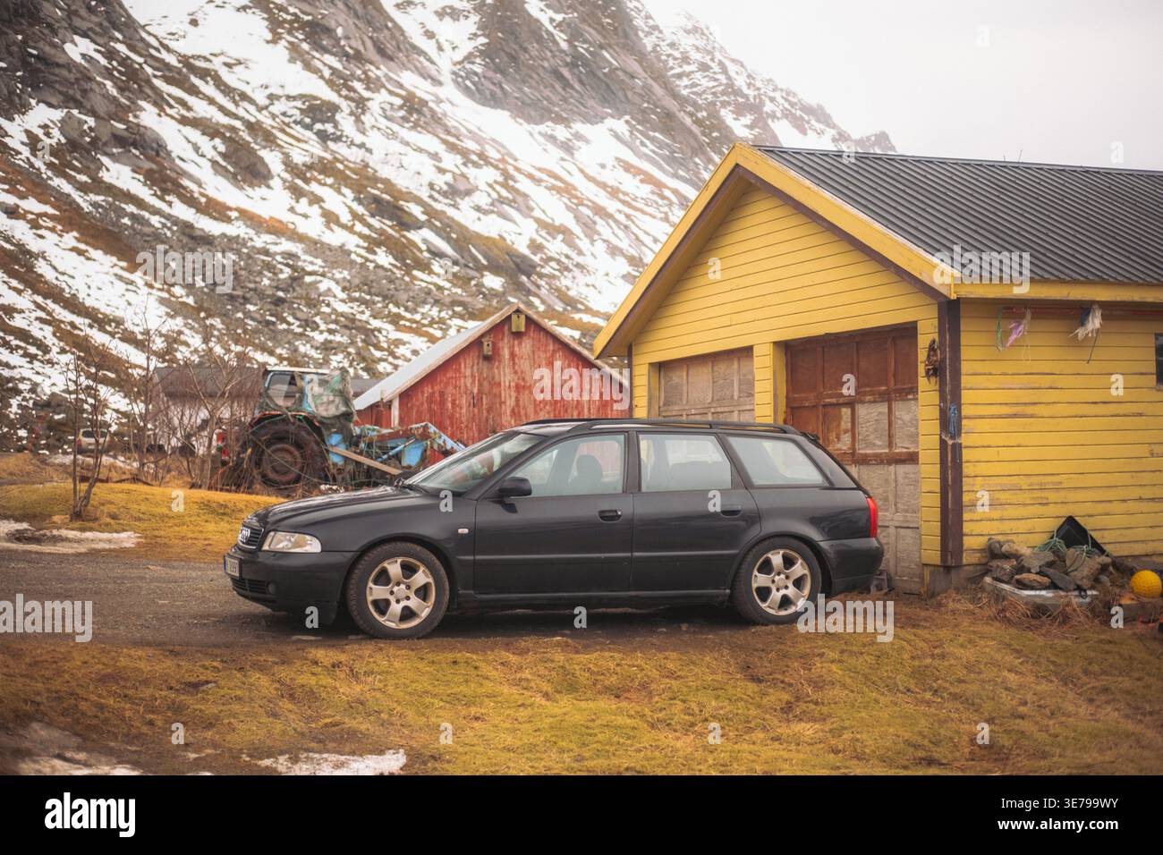 Blick auf einen schwarzen Kombi, der vor gelben und roten Holzgebäuden vor dem Hintergrund schneebedeckter Berge geparkt ist, Skjelfjord, Norwegen. Stockfoto