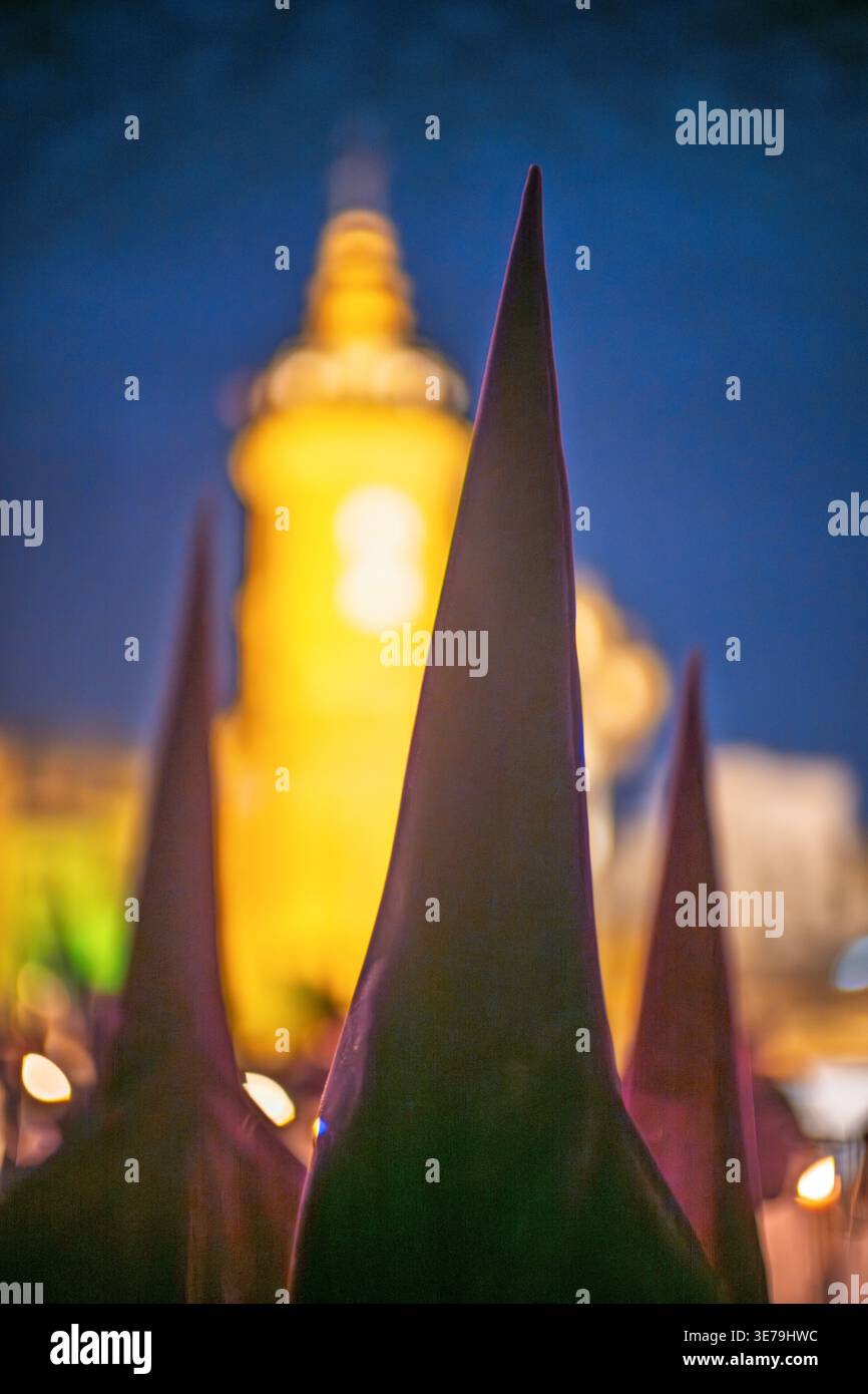 Die Nazarener der Bruderschaft La Estrella ziehen nachts während der Karwoche in Sevilla, Spanien. Konische Hauben im Vordergrund verschwimmen die Carmen Chapel. Stockfoto