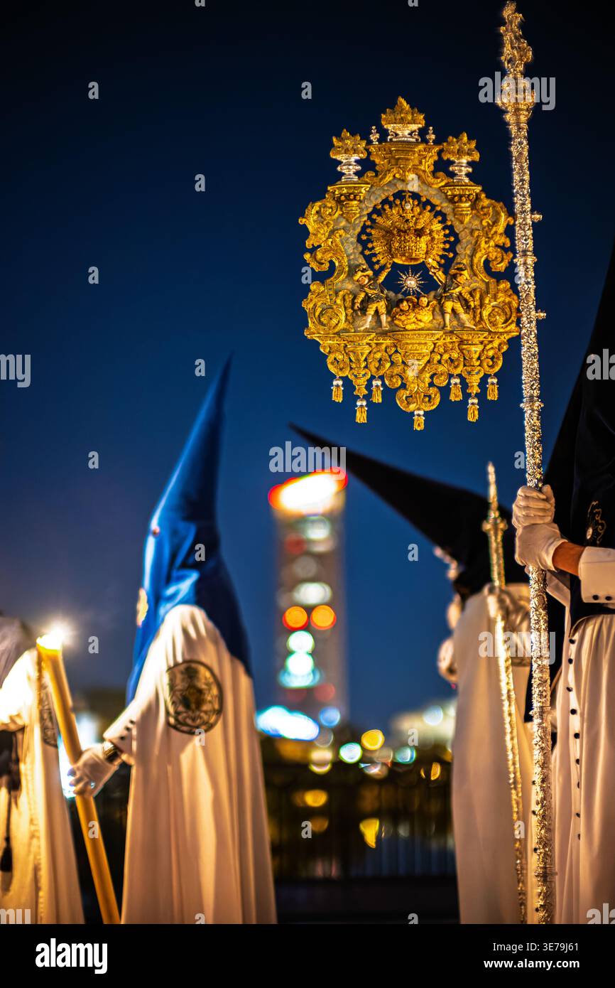 Ein Nazarener aus der Bruderschaft La Estrella hält ein Banner während einer Karwoche bei Nacht auf der Triana-Brücke in Sevilla. Wolkenkratzer Torre Sevilla Stockfoto