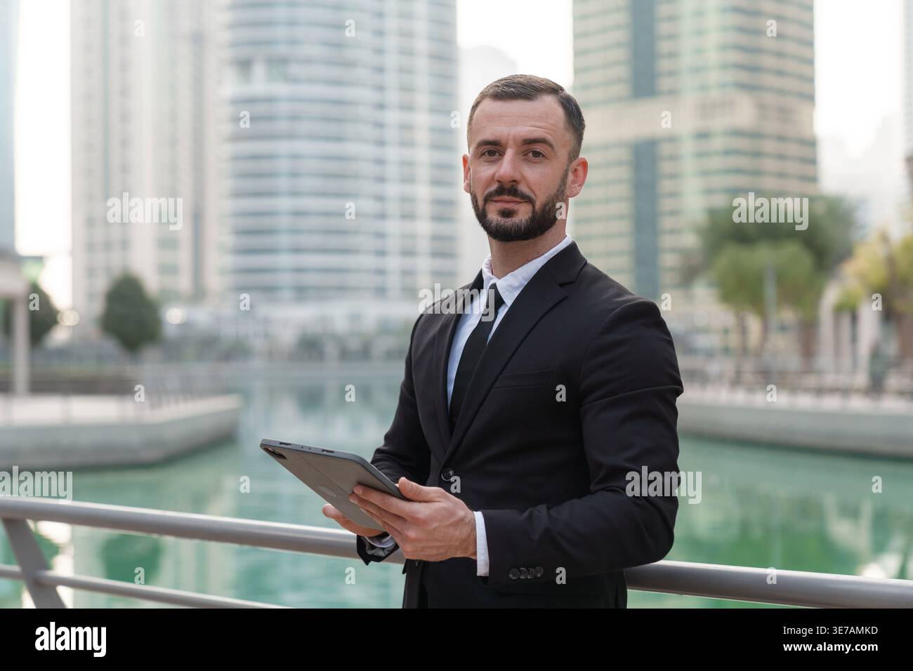 Geschäftsmann in formellen Anzügen steht auf der Terrasse am Wasser in Dubai Marina. Er benutzt ein Tablet, während die Skyline der Stadt dahinter auftaucht. Stockfoto