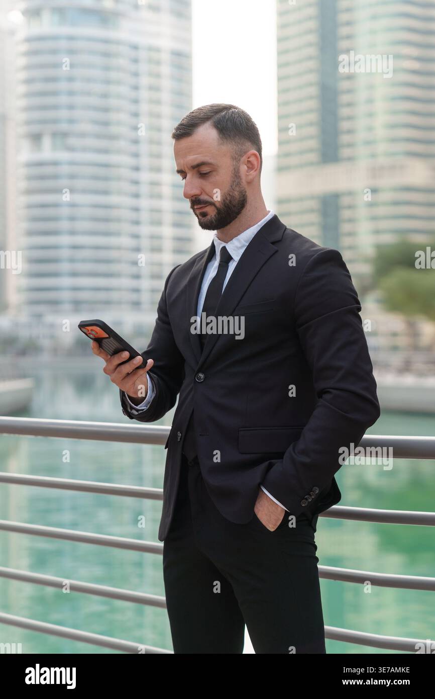 Geschäftsmann in formellem Anzug steht tagsüber am Geländer mit Smartphone und der Skyline von Dubai Marina im Hintergrund. Stockfoto