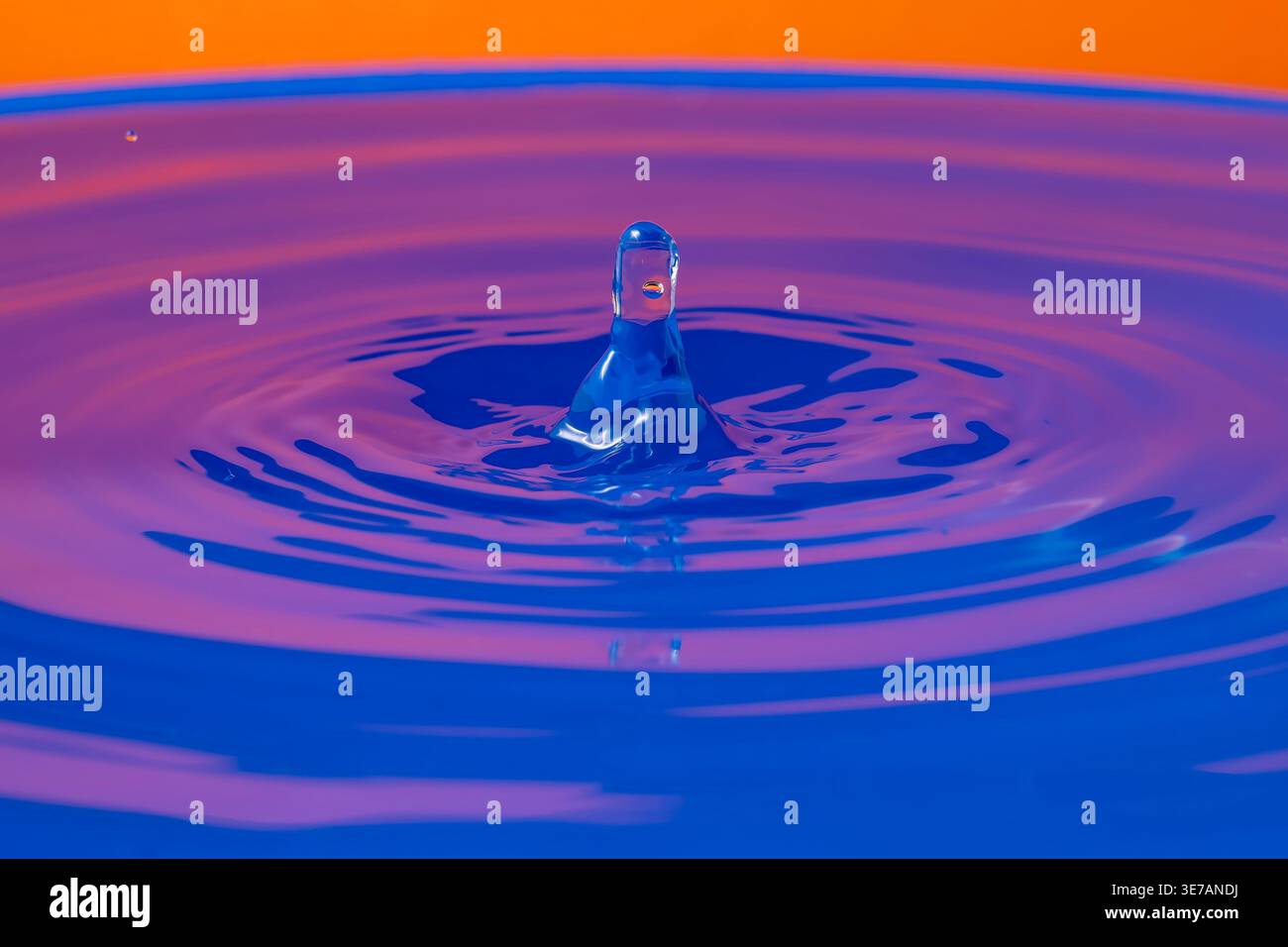 Ein Wassertropfen, der von oben fällt, prallt auf eine Wasseroberfläche und erzeugt einen grafischen Effekt auf einem blauen und orangen Hintergrund Stockfoto