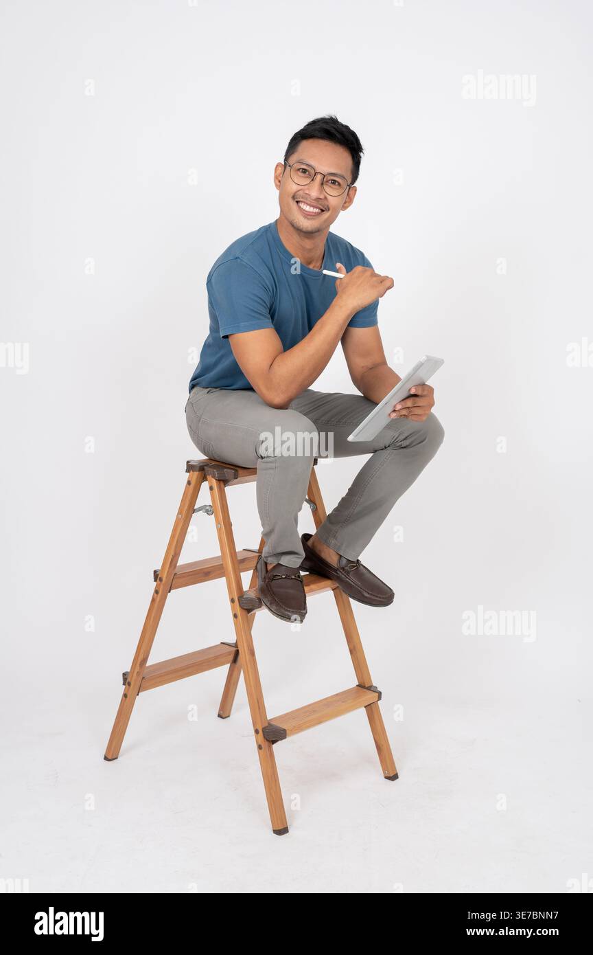 Lächelnder asiatischer Mann mit Stift, der Tablette hält, sitzt auf hölzerner Leiter vor weißem Hintergrund im Studio Stockfoto