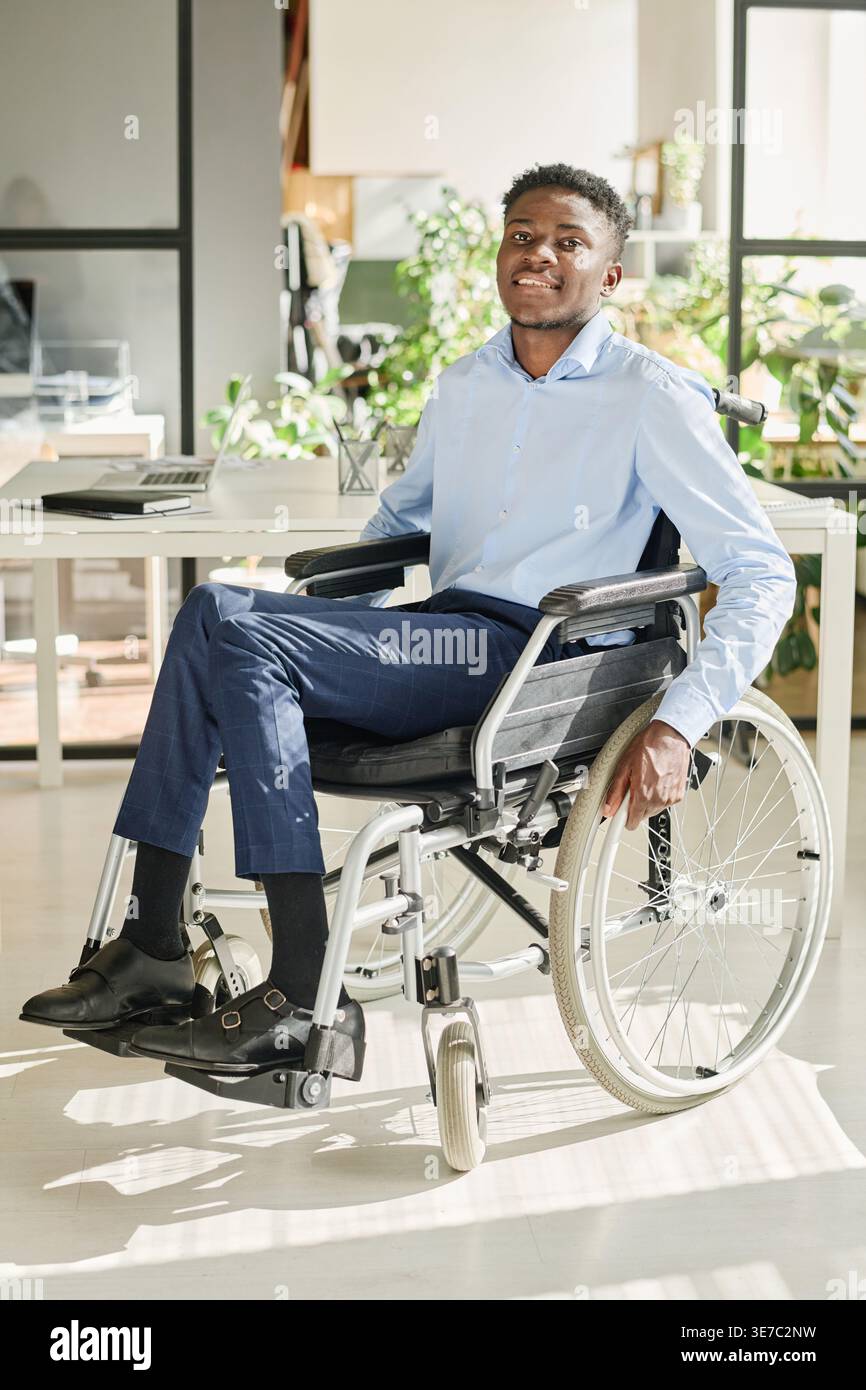 Lächelnder behinderter schwarzer Geschäftsmann, der während der Arbeit im Büro im Rollstuhl sitzt Stockfoto