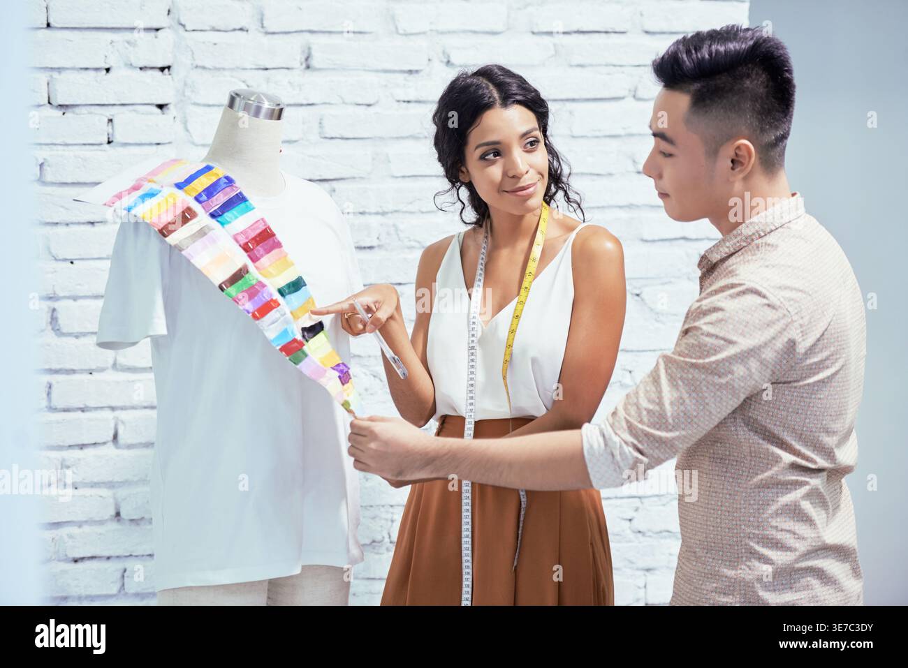 Hispanische und asiatische Modedesigner entscheiden sich für Stoffe, die im Werkstattstudio an einer Schaufensterpuppe mit Hemd an der Ziegelwand stehen Stockfoto