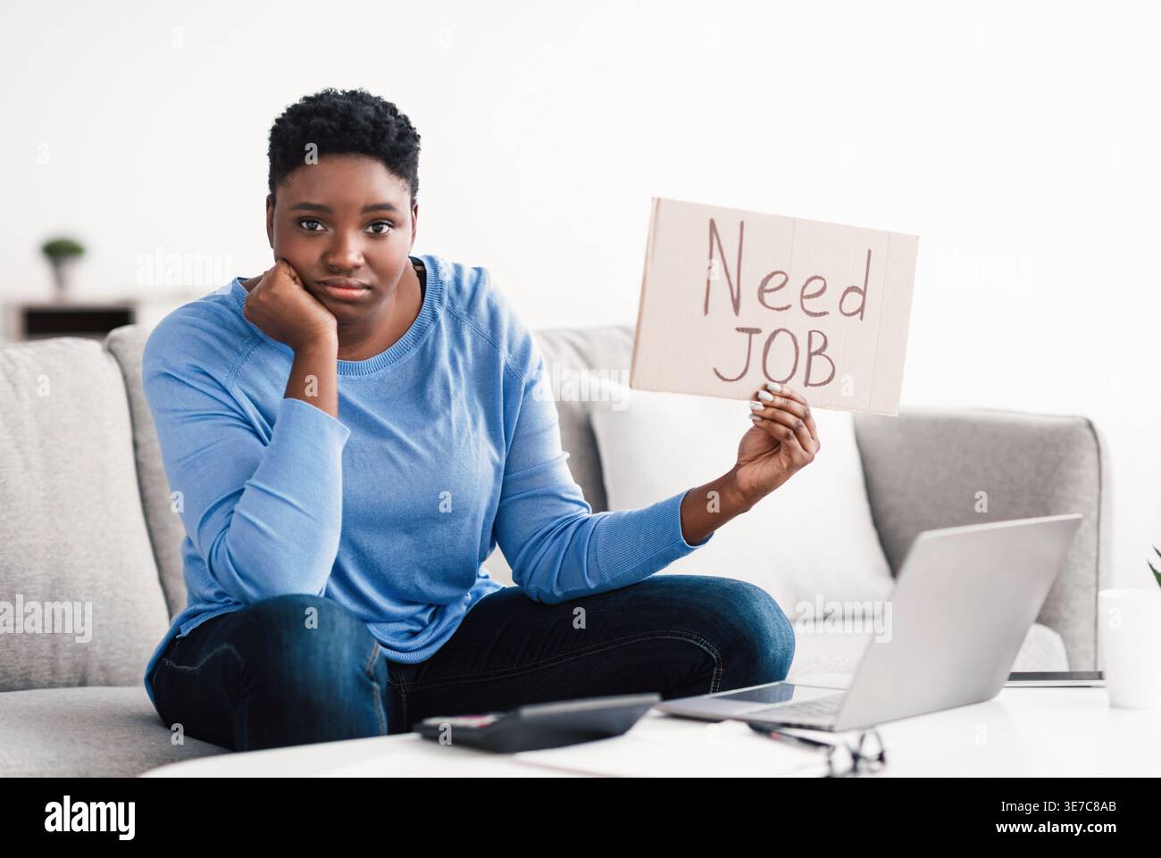 Schwarze Frau mit kurzem Afro-Schild mit Schreibtext, der auf der Couch sitzt und im Wohnzimmer zu Hause den Kopf auf der Hand beugt Stockfoto