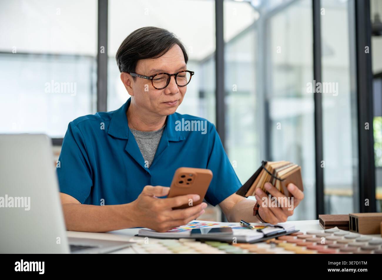 Asiatischer männlicher Innenarchitekt mittleren Alters mit smLifestylephone mit Holzproben am Tisch im Büro Stockfoto