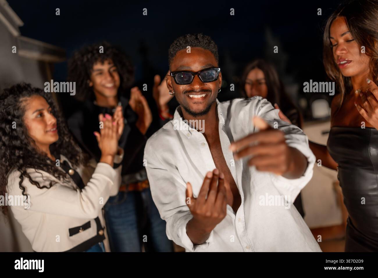Schwarzer Mann mit Sonnenbrille und weißem Hemd, der nachts mit Freunden auf dem Dach tanzt Stockfoto