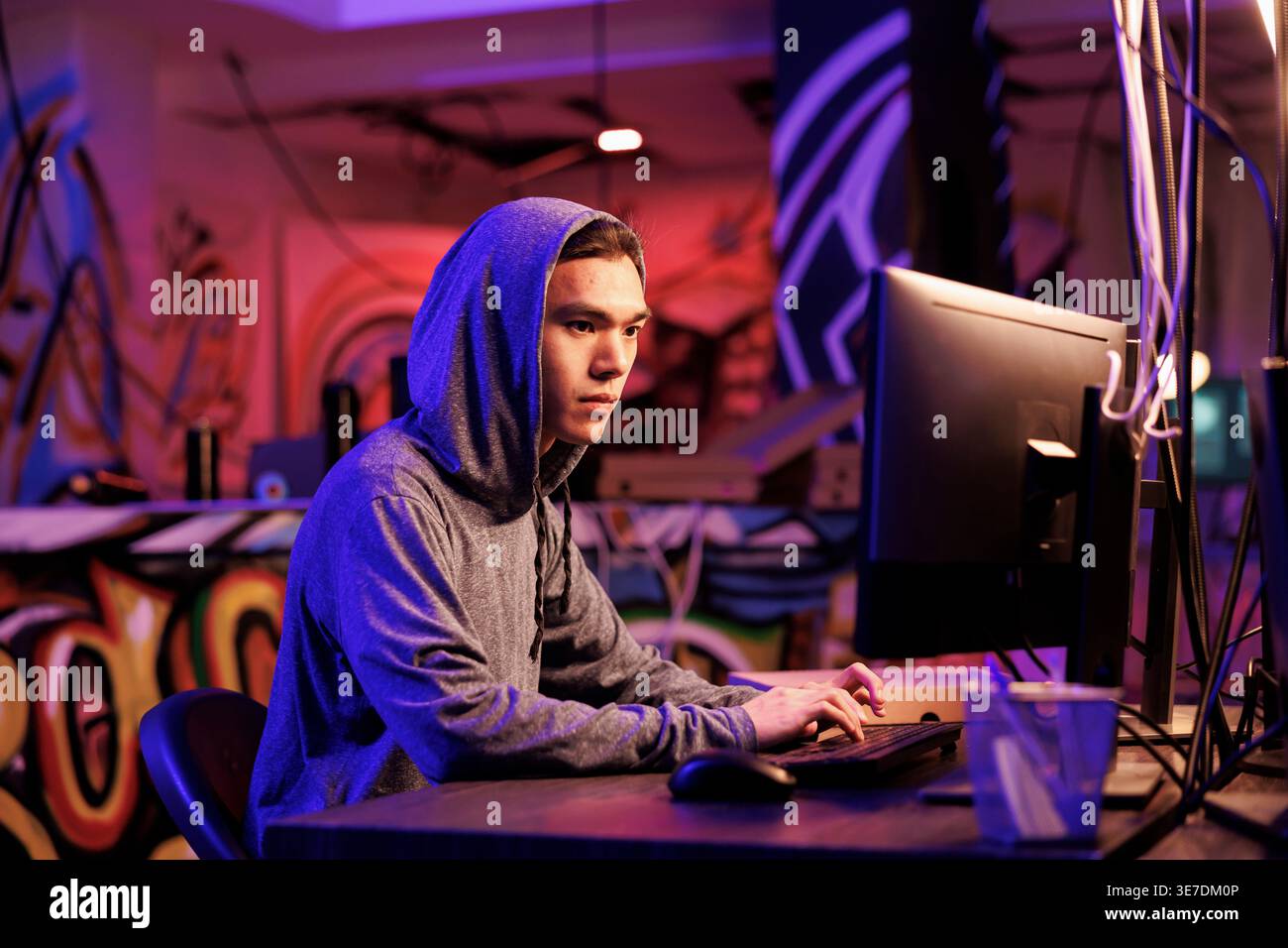 Asiatischer Internet-Betrüger knackt Passwort auf dem Computer, um Zugriff auf die Datenbank zu erhalten. Junger Mann in der Kapuze, der schädlichen Software-Code auf dem Computer schreibt und in einem dunklen Raum in den Server einbricht Stockfoto