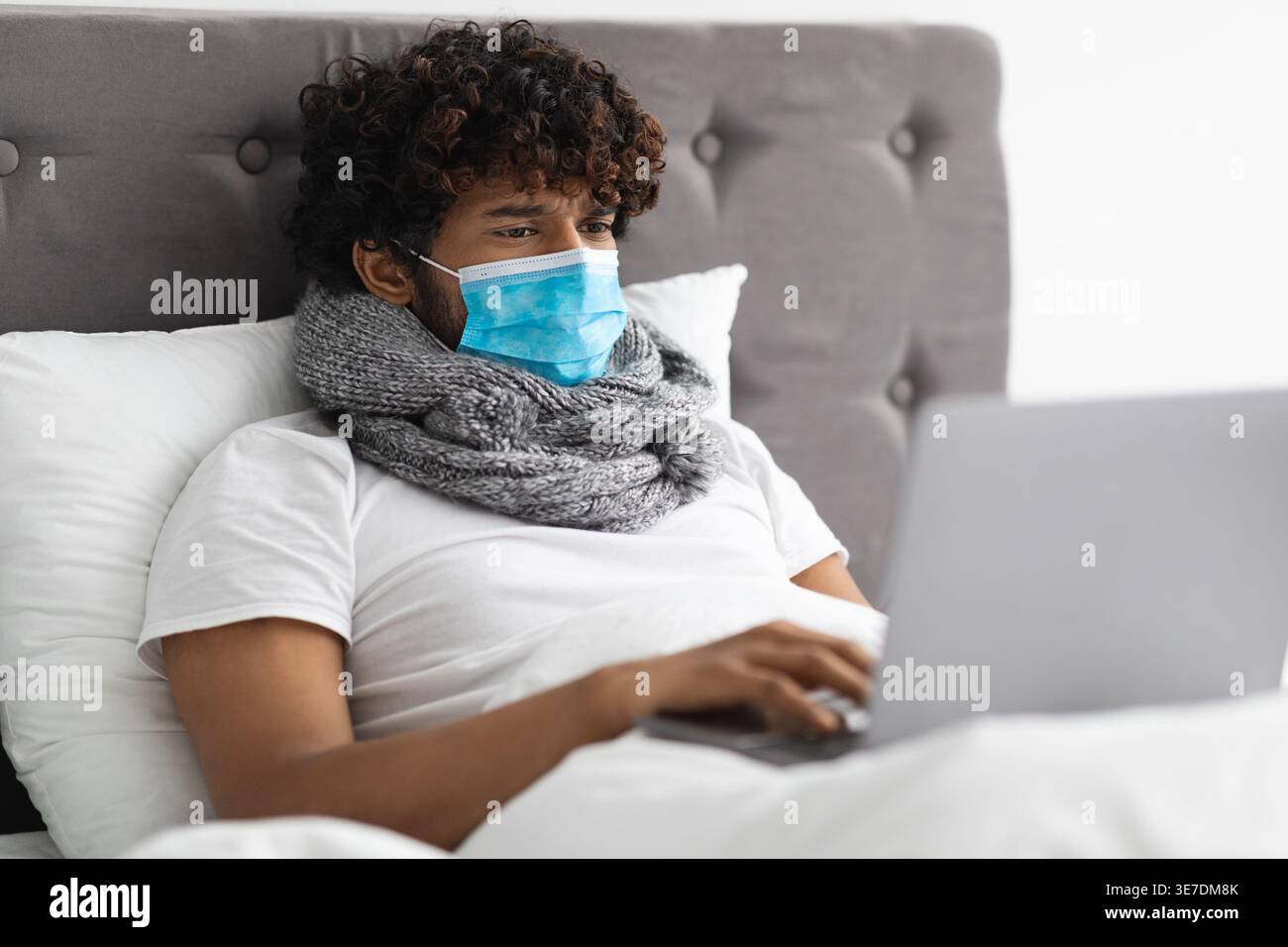 Kranker asiatischer Mann mit lockigem Haar, der Schal um den Hals und Gesichtsmaske mit Laptop trägt, während er zu Hause im Bett lag Stockfoto