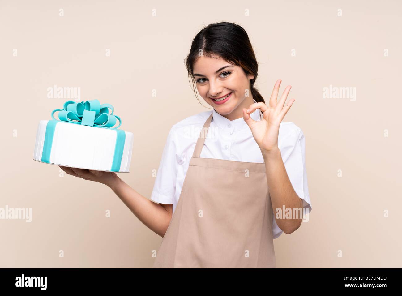 Hispanische Konditorin mit OK-Hand-Schild mit Kuchen, der vor beigem Hintergrund im Studio steht Stockfoto