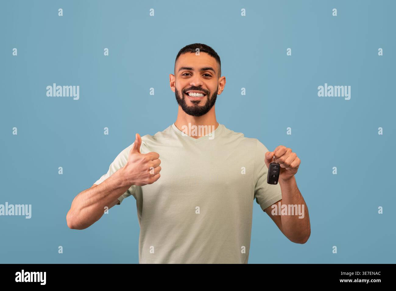 Ein lächelnder Mann aus dem Nahen Osten mit Bart, der den Autoschlüssel hält und die Daumen vor blauem Hintergrund im Studio zeigt Stockfoto