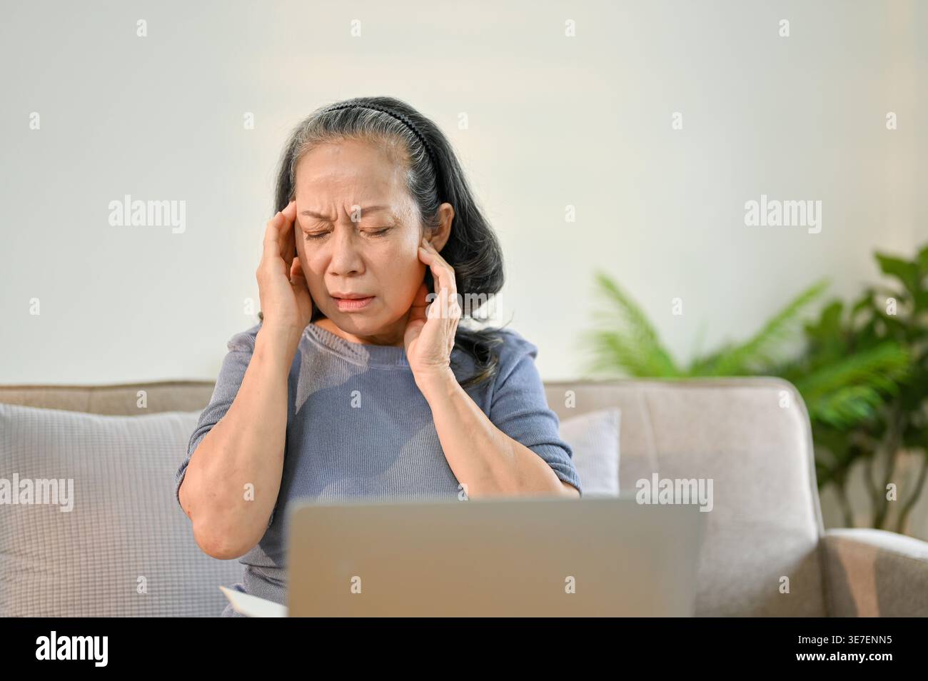 Asiatische Seniorin, die ihre Schläfe berührt und Kopfschmerzen hat, sitzend auf dem Sofa mit Laptop zu Hause Stockfoto