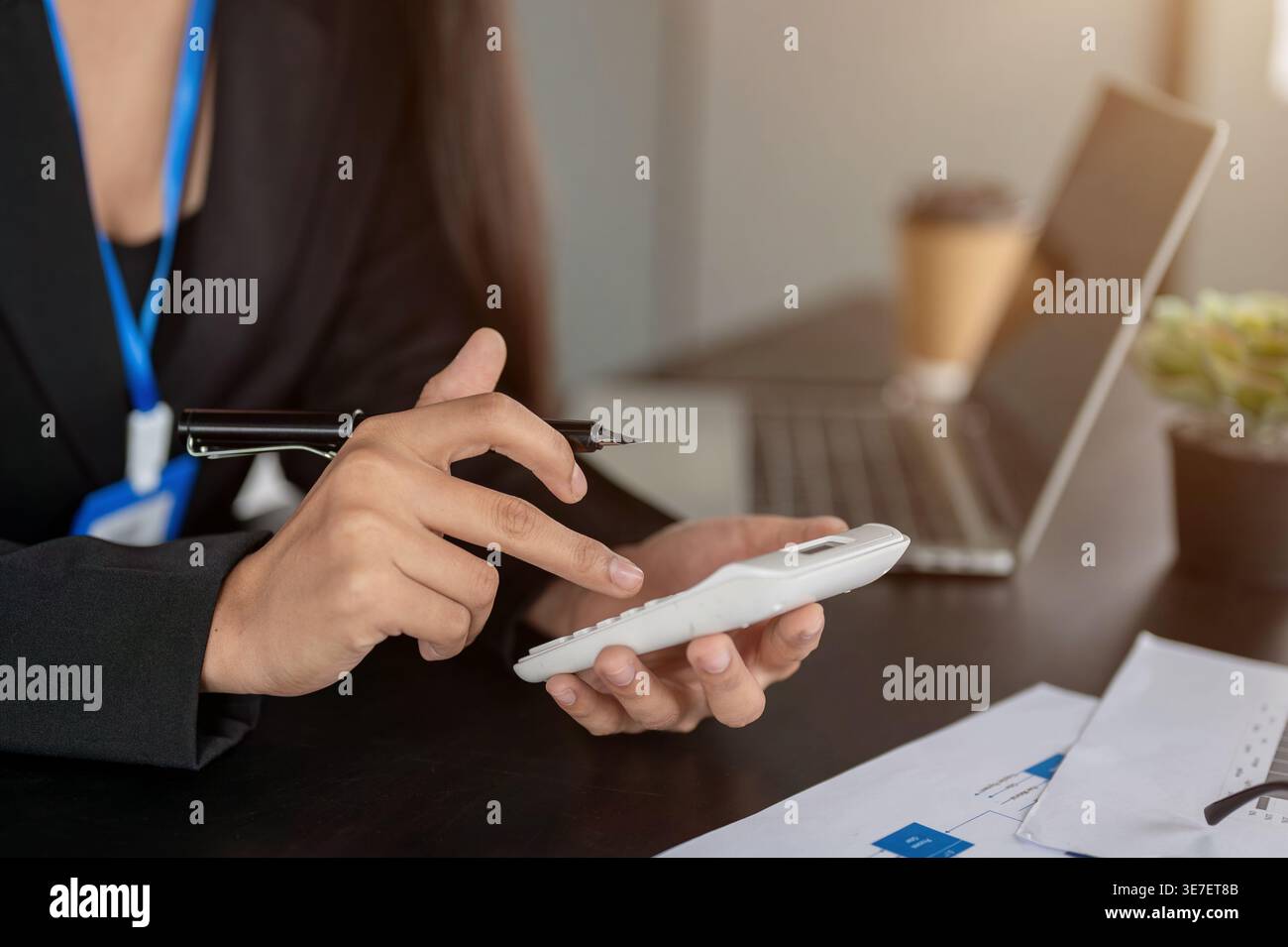 Asiatische Geschäftsfrau hält Stift und benutzt Taschenrechner, während sie im Büro mit Laptop und Dokumenten an ihrem Schreibtisch sitzt Stockfoto