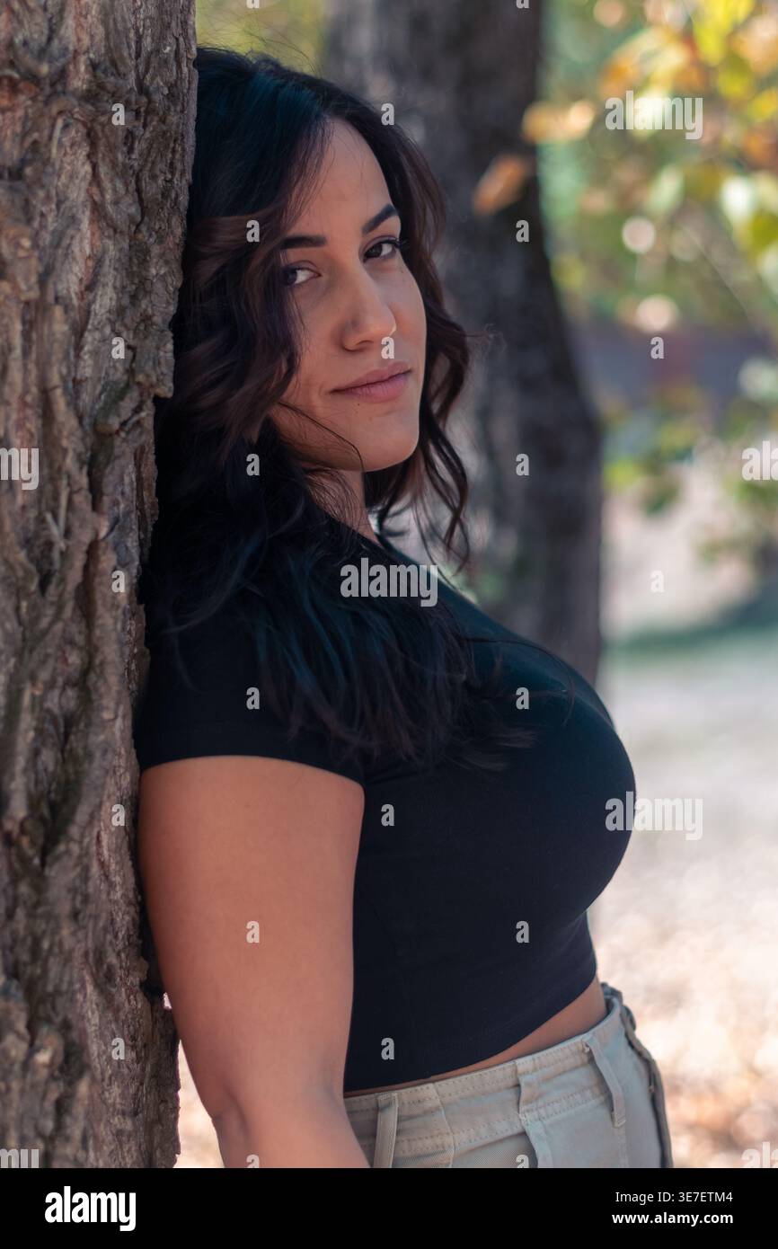 Ernste hispanische Frau in schwarzem Hemd, die tagsüber im Park an einem Baum steht und sich lehnt Stockfoto