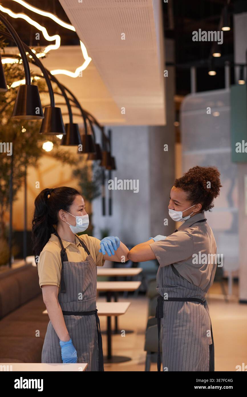 Gemischte und asiatische Kellnerinnen mit Gesichtsmasken und Schürzen, die sich gegenseitig am Ellenbogen stoßen, während sie während der Pandemie im Café stehen Stockfoto