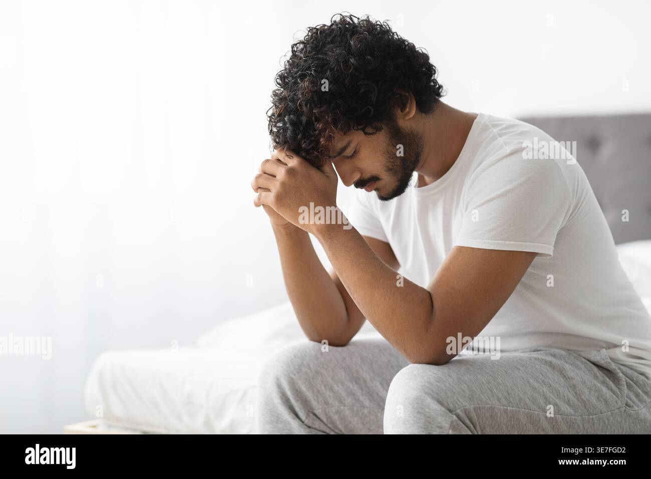 Gestresster bärtiger indischer Mann mit lockigem Haar, der die Stirn auf geballten Fäusten lehnt und im Schlafzimmer zu Hause auf dem Bett sitzt Stockfoto
