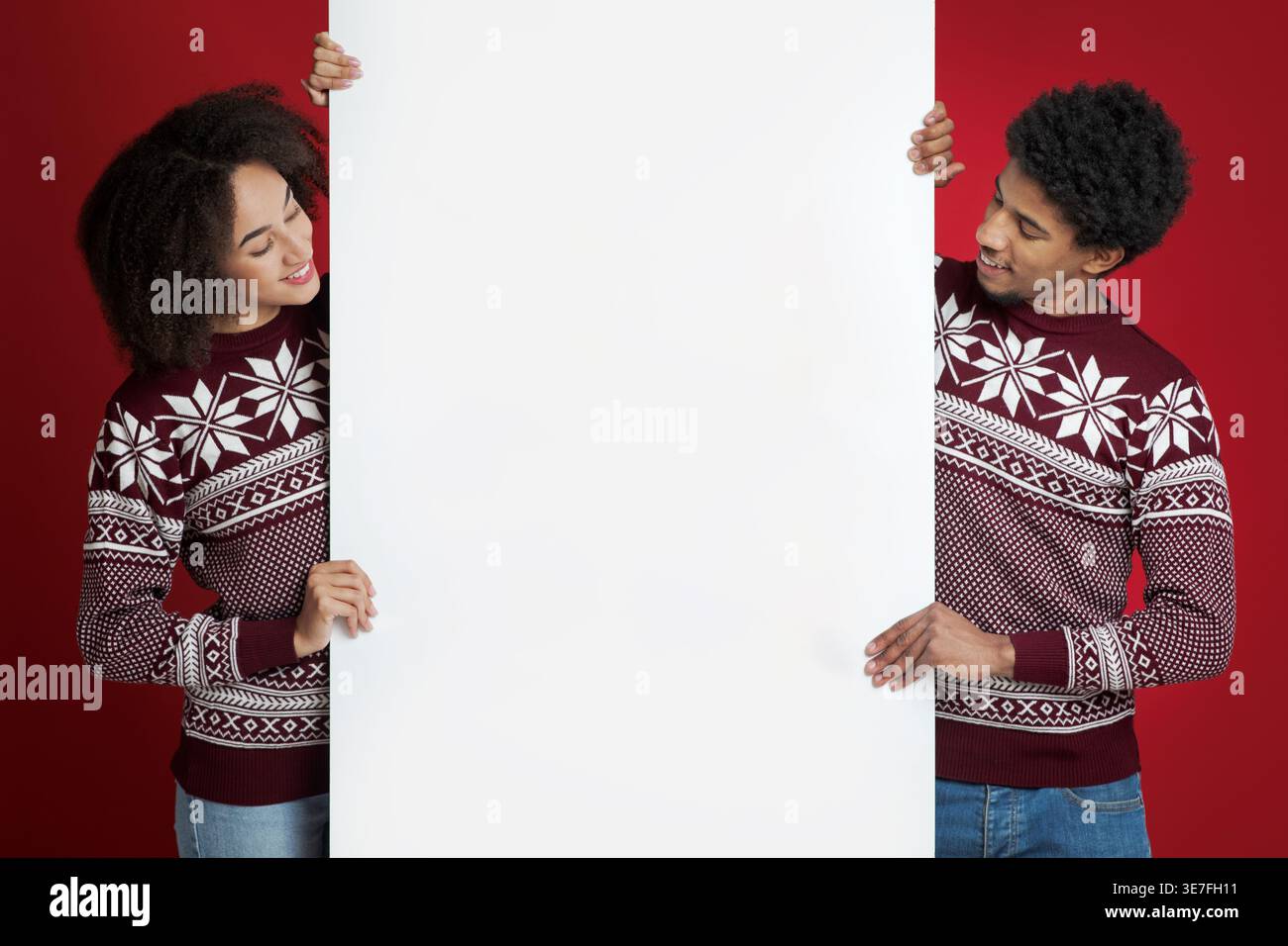 Schwarzes Paar mit Afro, der einen roten Pullover trägt und das weiße Banner zusammen vor rotem Hintergrund im Studio ansieht Stockfoto