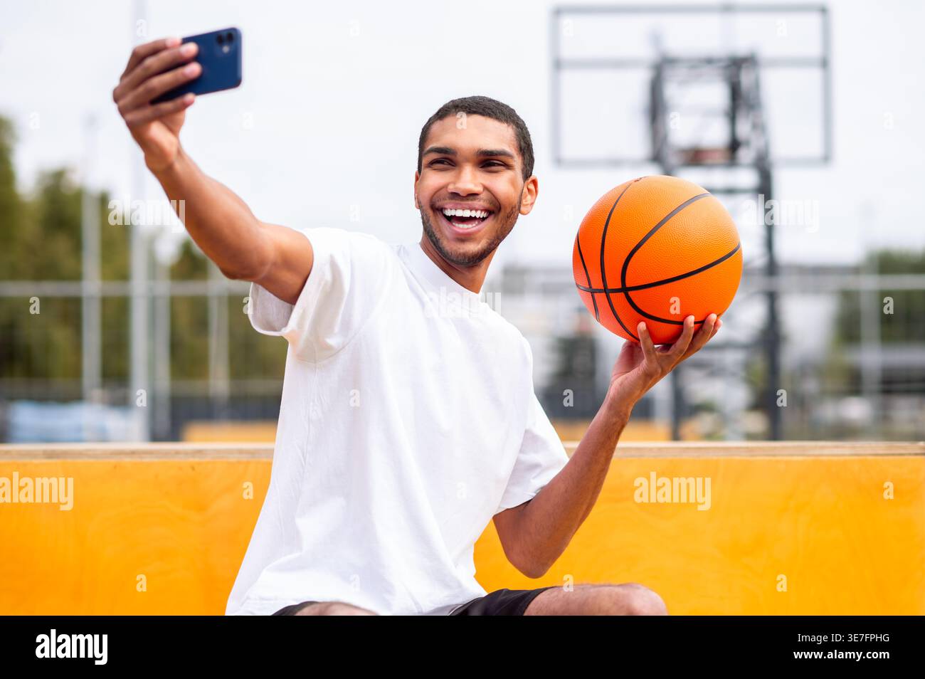 Lächelnder Schwarzer Mann, der tagsüber auf Beton sitzend im Basketballfeld im Park Selfie macht Stockfoto
