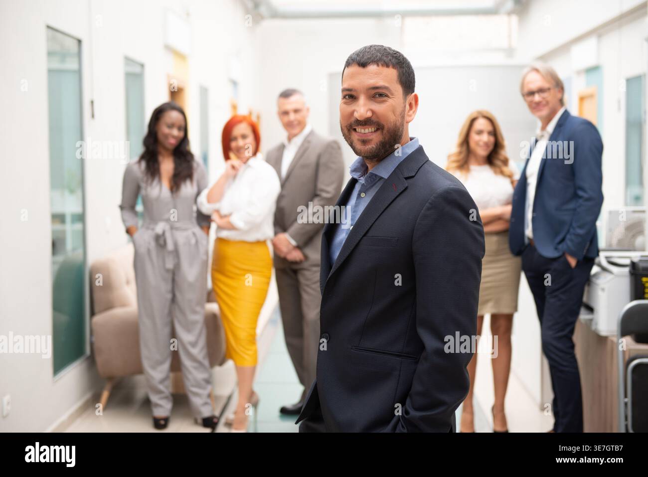 Lächelnder hispanischer Geschäftsmann, der mit Händen in Taschen in der Nähe verschiedener Kollegen im Büro steht Stockfoto