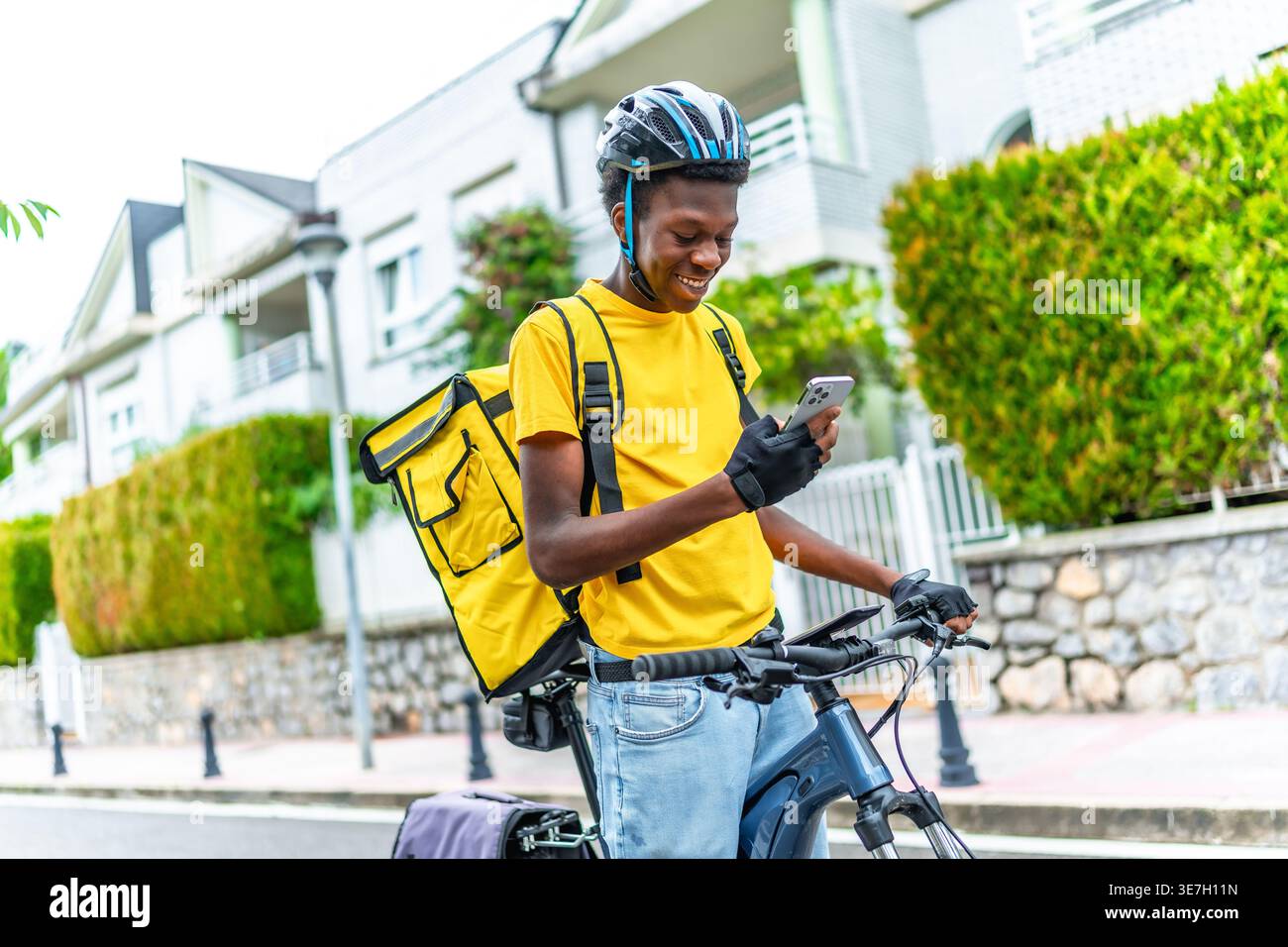 Schwarzer Mann, der Helm und Liefertasche trägt und das smLifestylephone benutzt, um den Kunden zu kontaktieren, während er tagsüber mit dem Fahrrad auf der Straße steht Stockfoto