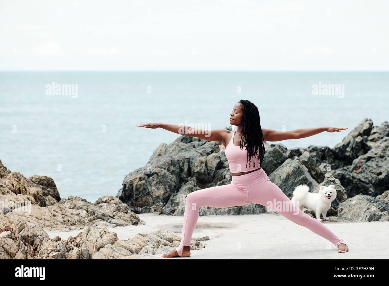 Schwarze Frau mit Dreadlocks im Fitness-Outfit, die Arme und Beine am Meer dehnt Stockfoto