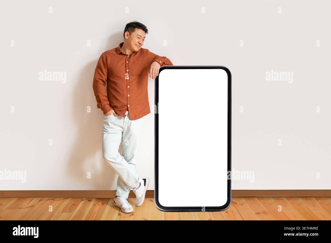 Asiatischer Mann, der Arm an einem großen Smartphone mit weißem Bildschirm lehnt, während er mit der Hand in der Tasche steht und die Beine zu Hause gegen die weiße Wand gekreuzt sind Stockfoto
