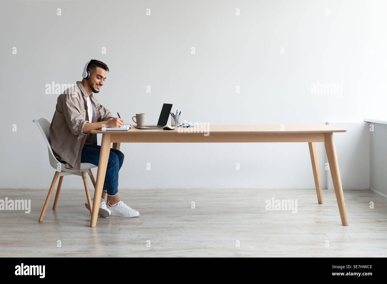 Mann aus dem Nahen Osten, der Kopfhörer trägt und Notizen schreibt, während er an einem Laptop arbeitet und am Tisch im Büro sitzt Stockfoto