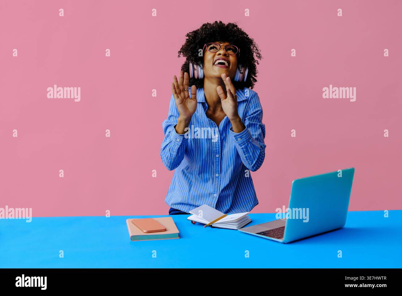 Schwarze Geschäftsfrau mit lockigem Haar, die eine Brille trägt, Musik über Kopfhörer hört und aufblickt, während sie am Tisch mit Laptop gegen pi steht... Stockfoto