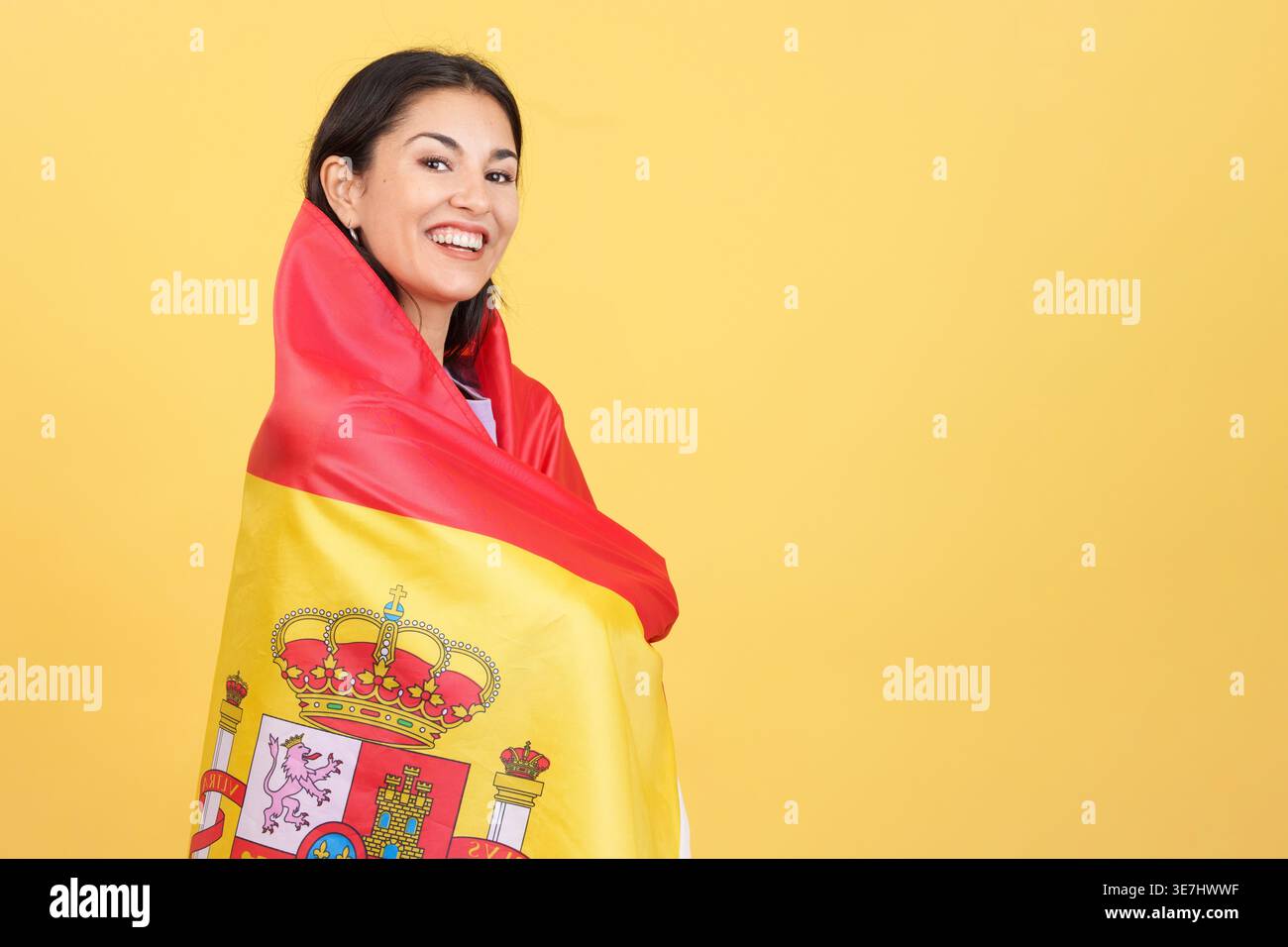 Lächelnde hispanische Frau, die in die spanische Nationalflagge gewickelt ist und im Studio auf gelbem Hintergrund steht Stockfoto
