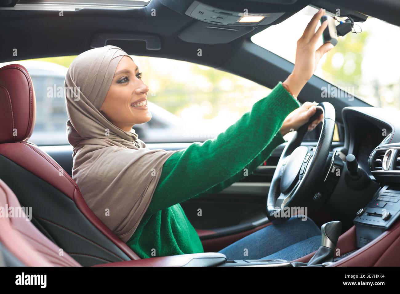 Lächelnde Frau aus dem Nahen Osten, die tagsüber einen Hijab trägt, der den Rückspiegel beim Autofahren fixiert Stockfoto