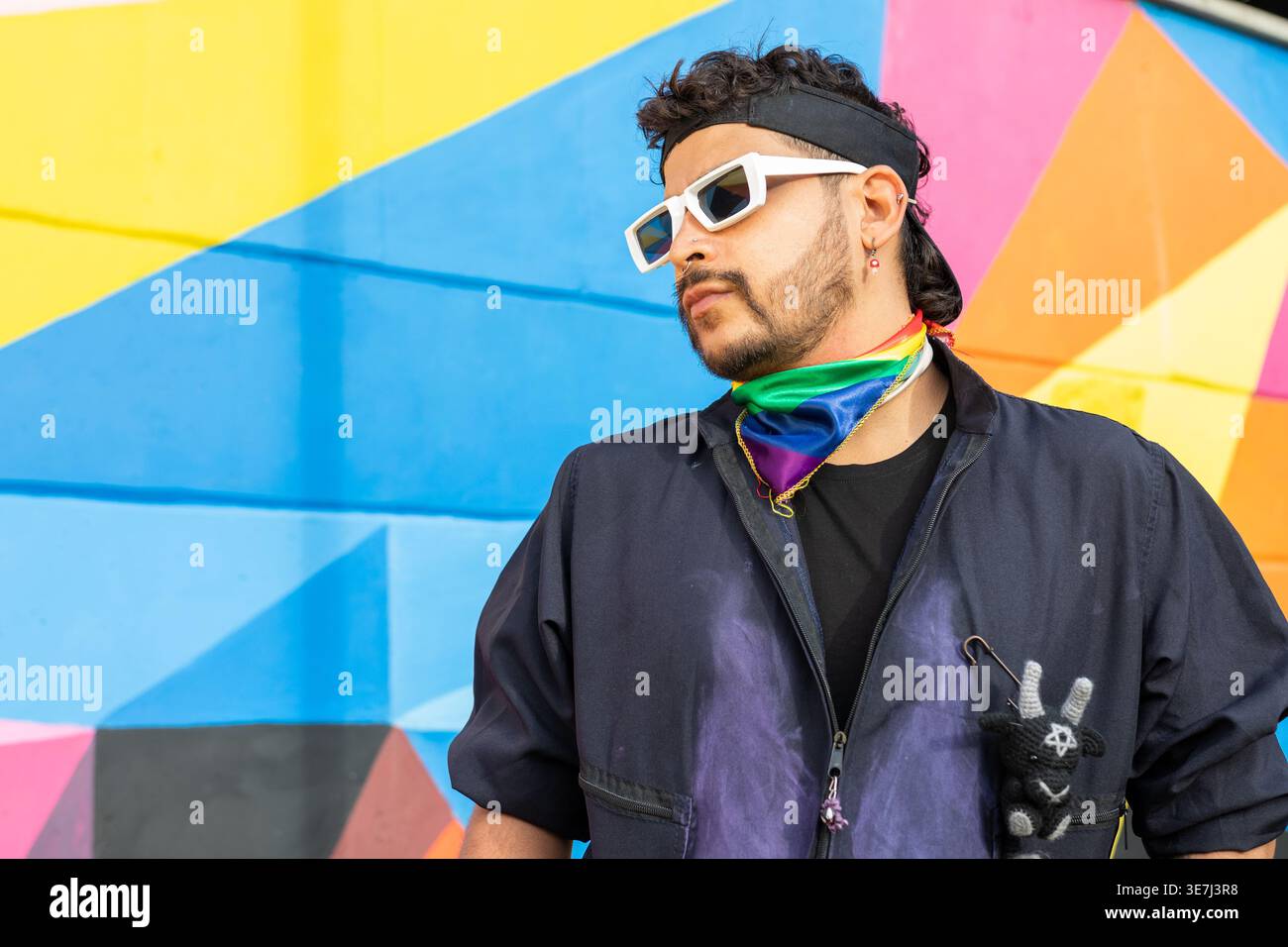 Hispanischer schwuler Mann in Sonnenbrille mit Stolz-Bandana, der im Park vor einer bunten Wand steht Stockfoto