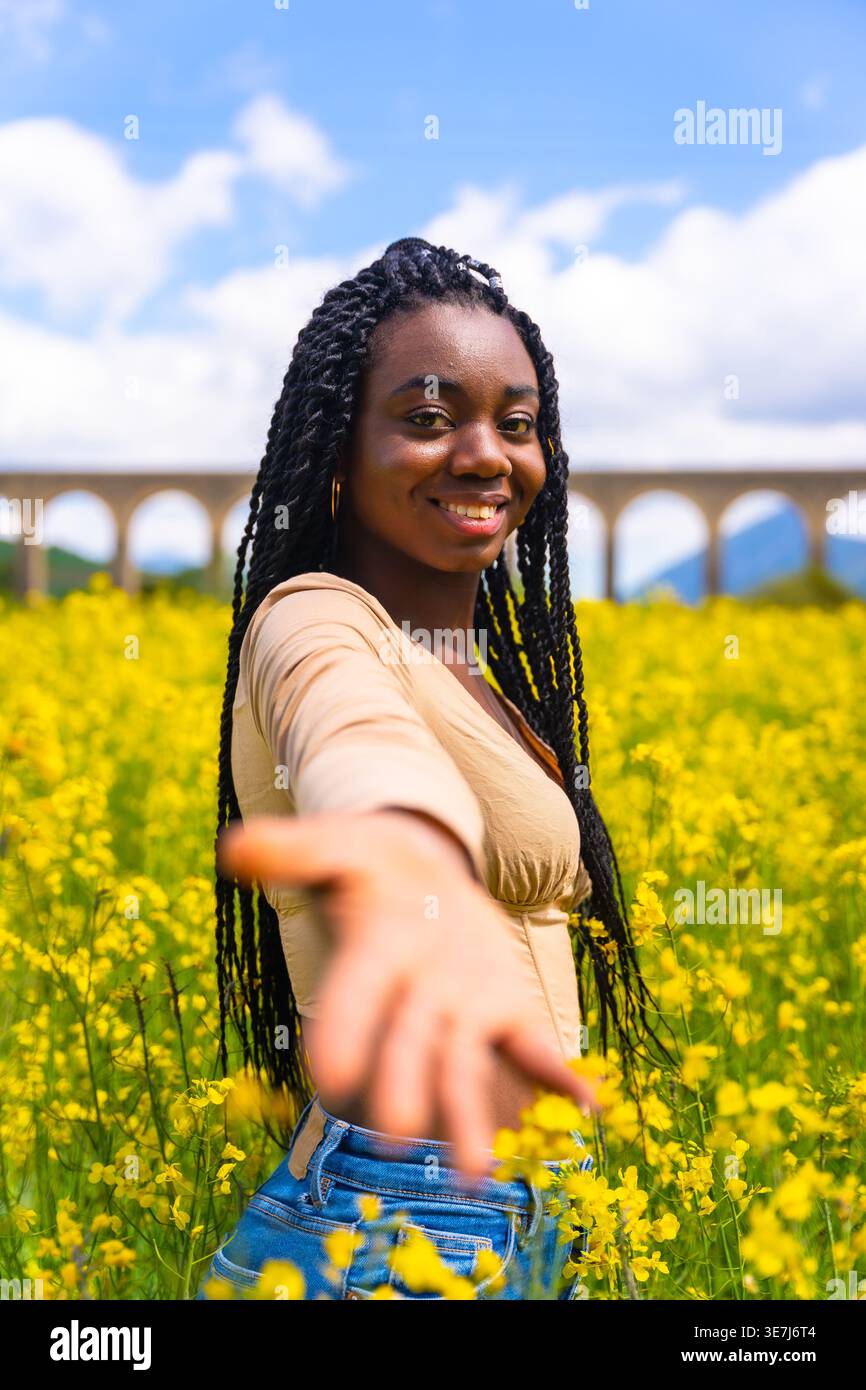 Lächelnde schwarze Frau mit Wendungen, die mit offenen Händen im Feld der gelben Blumen an sonnigem Tag steht Stockfoto