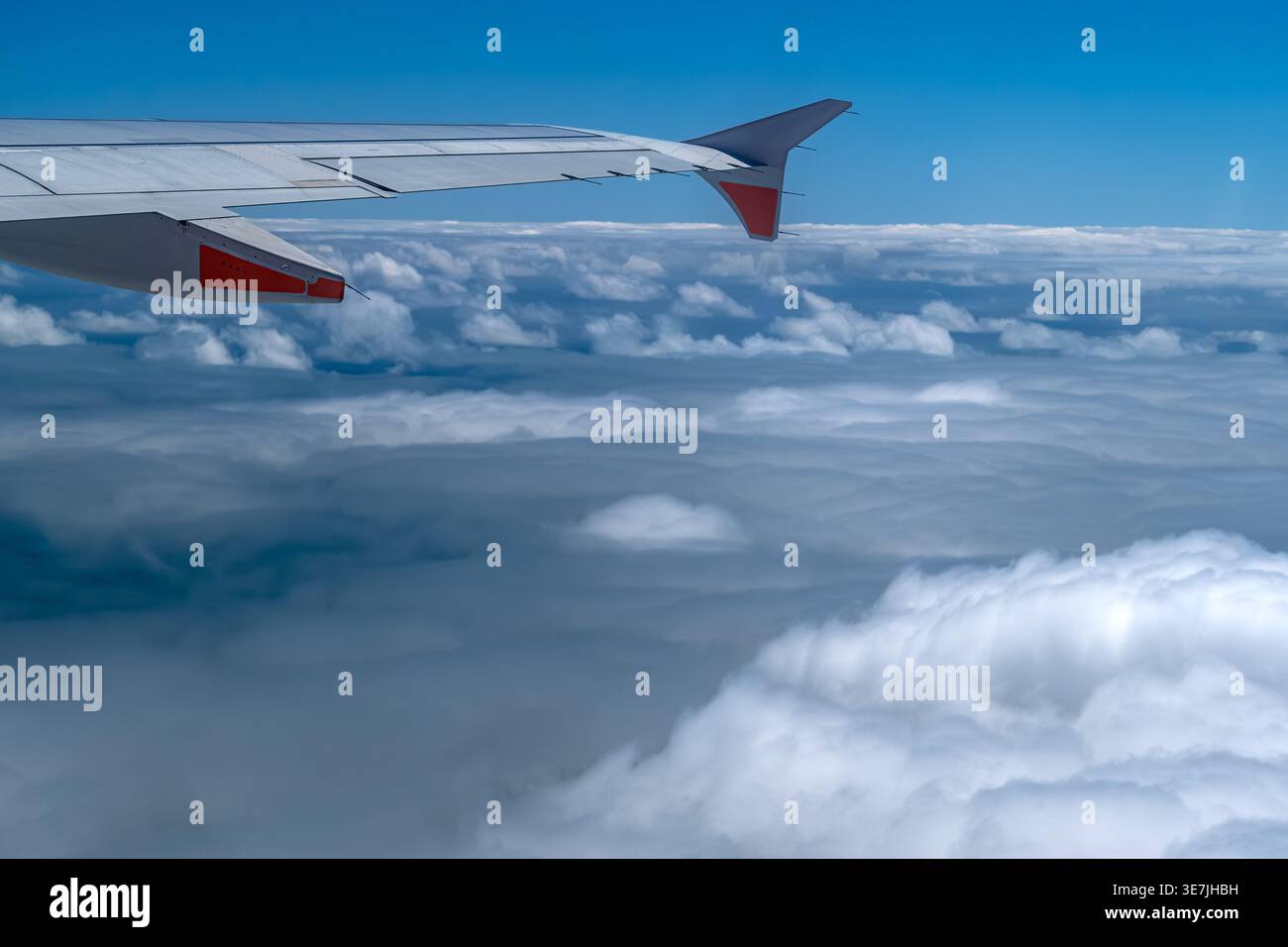 Blick von einem Passagierflugzeugfenster aus, das den Flugzeugflügel mit roten Akzenten zeigt, der über einer dramatischen Schicht weißer Cumulus-Wolken unter einer lebendigen Decke aufragt Stockfoto