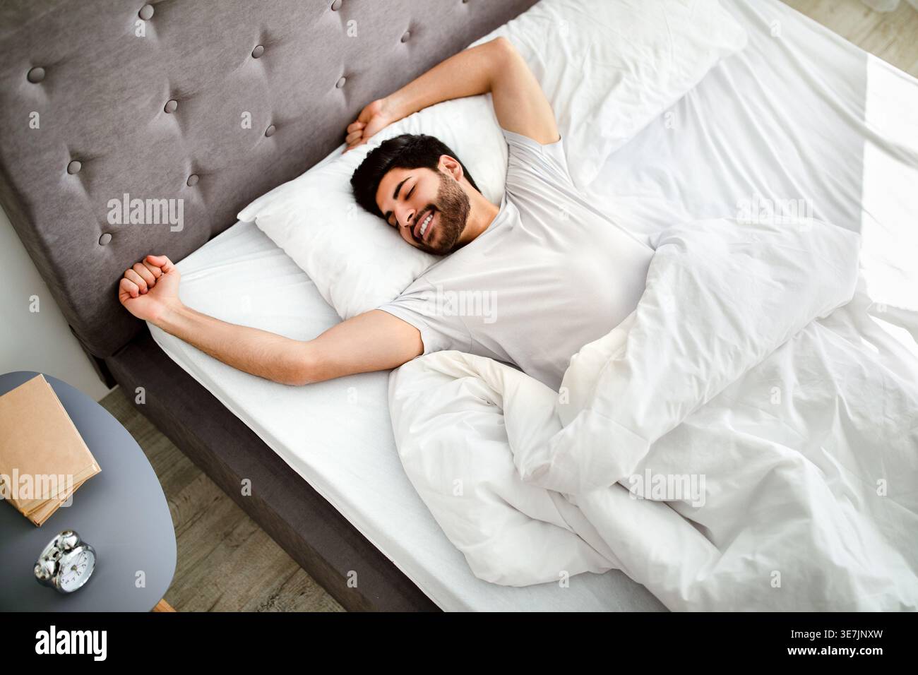 Ein arabischer Mann mit Bart, der ein weißes Hemd trägt und die Arme mit geschlossenen Augen ausdehnt, während er zu Hause auf dem Bett liegt Stockfoto