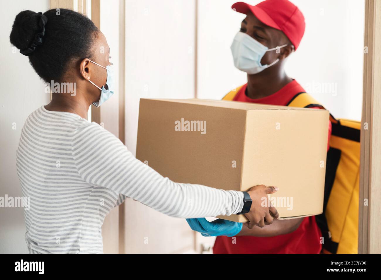 Schwarzer Liefermann mit Gesichtsmaske, der den Paketkasten an weibliche Kunden übergibt, während er während der Pandemie vor der Haustür zu Hause steht Stockfoto