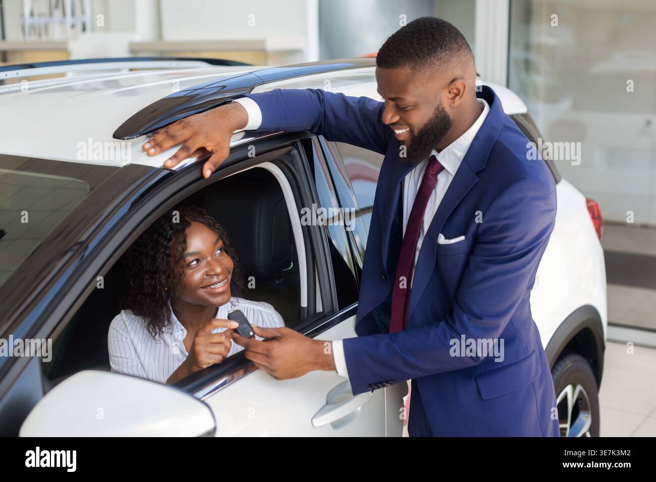 Schwarzer Verkäufer im Business-Anzug, der weiblichen Kundin, die im Auto sitzt, Autoschlüssel gibt Stockfoto