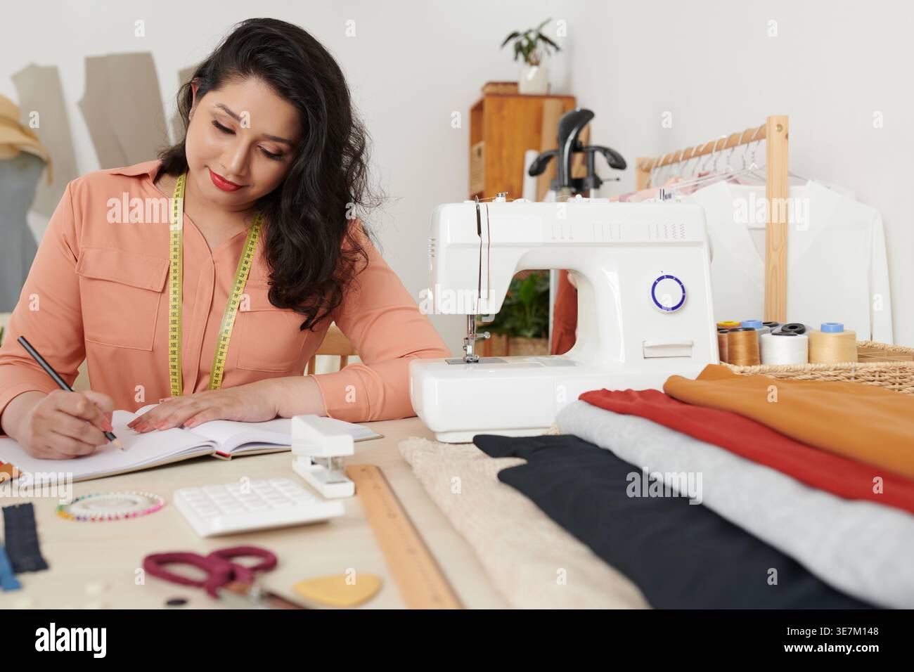 Lächelnde asiatische Schneiderin, die Notizen schreibt und mit Nähmaschine in der Werkstatt am Tisch sitzt Stockfoto