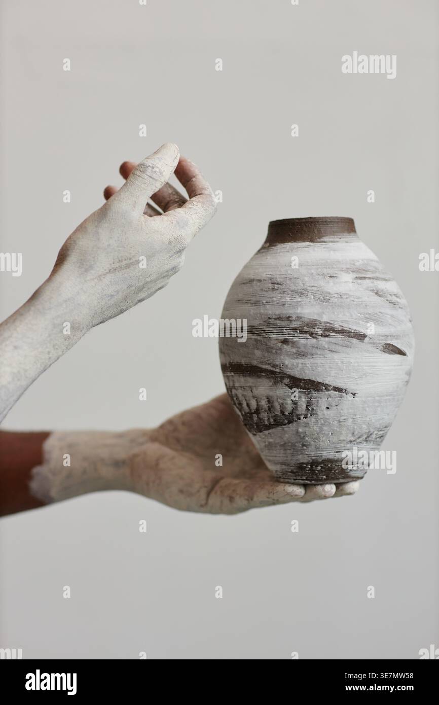Zugeschnittenes Bild eines schwarzen männlichen Künstlers, der handgefertigte Keramikvase hält, die mit farbigen Händen und Armen im Tonerstudio steht Stockfoto