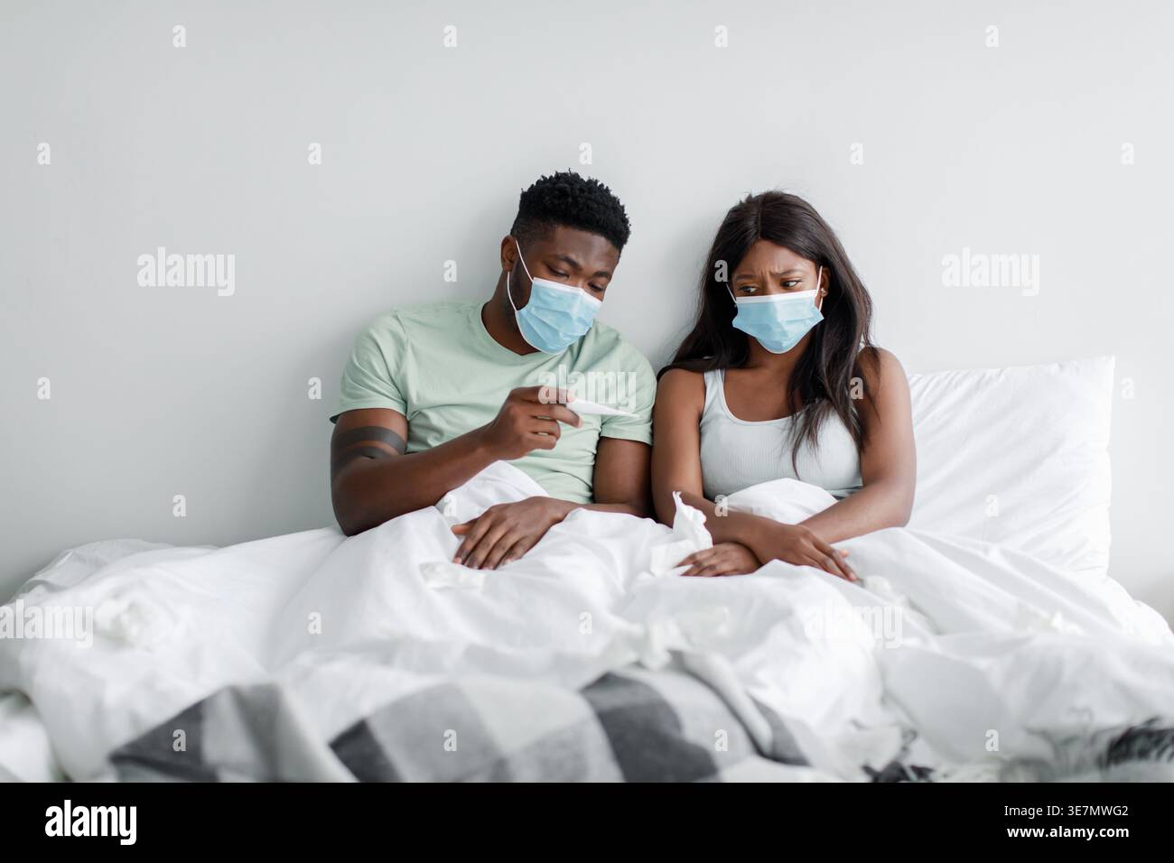 Schwarzes Paar, das Gesichtsmasken trägt und auf das Thermometer blickt, während es zu Hause auf dem Bett sitzt Stockfoto
