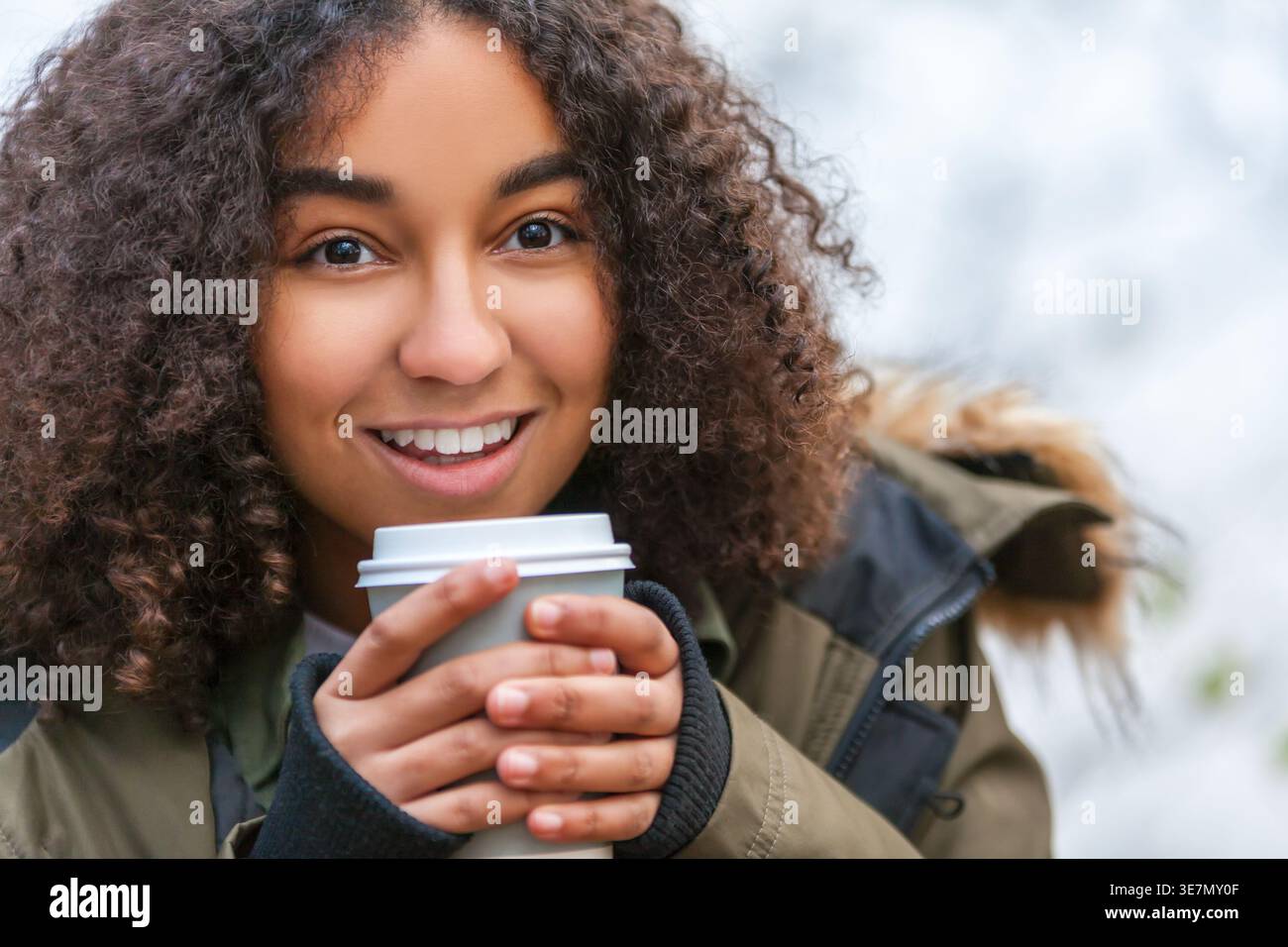 Lächelnde junge Frau mit lockigen Haaren, die warme Winterjacke trägt und im Freien Kaffee hält Stockfoto