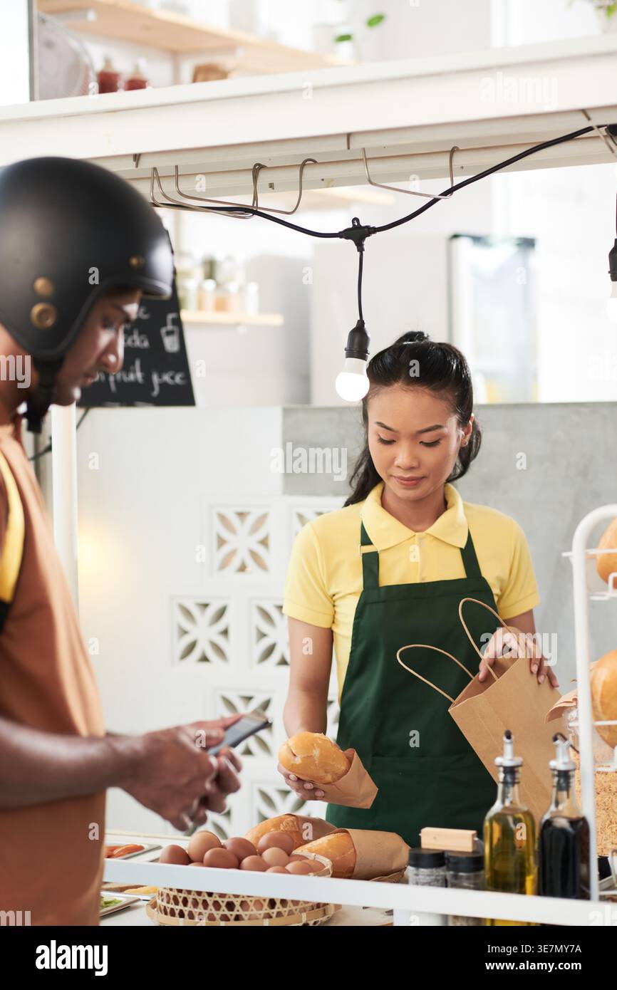 Asiatische Arbeitnehmerin, die verpackte Sandwiches in Papiertüte zum Mitnehmen im Laden verpackt Stockfoto