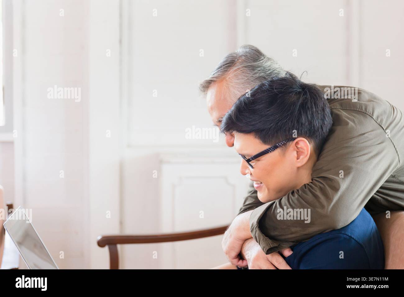 Asiatischer älterer Vater, der hinter ihm steht und einen Mann mittleren Alters umarmt, indem er zu Hause auf der Couch sitzt Stockfoto